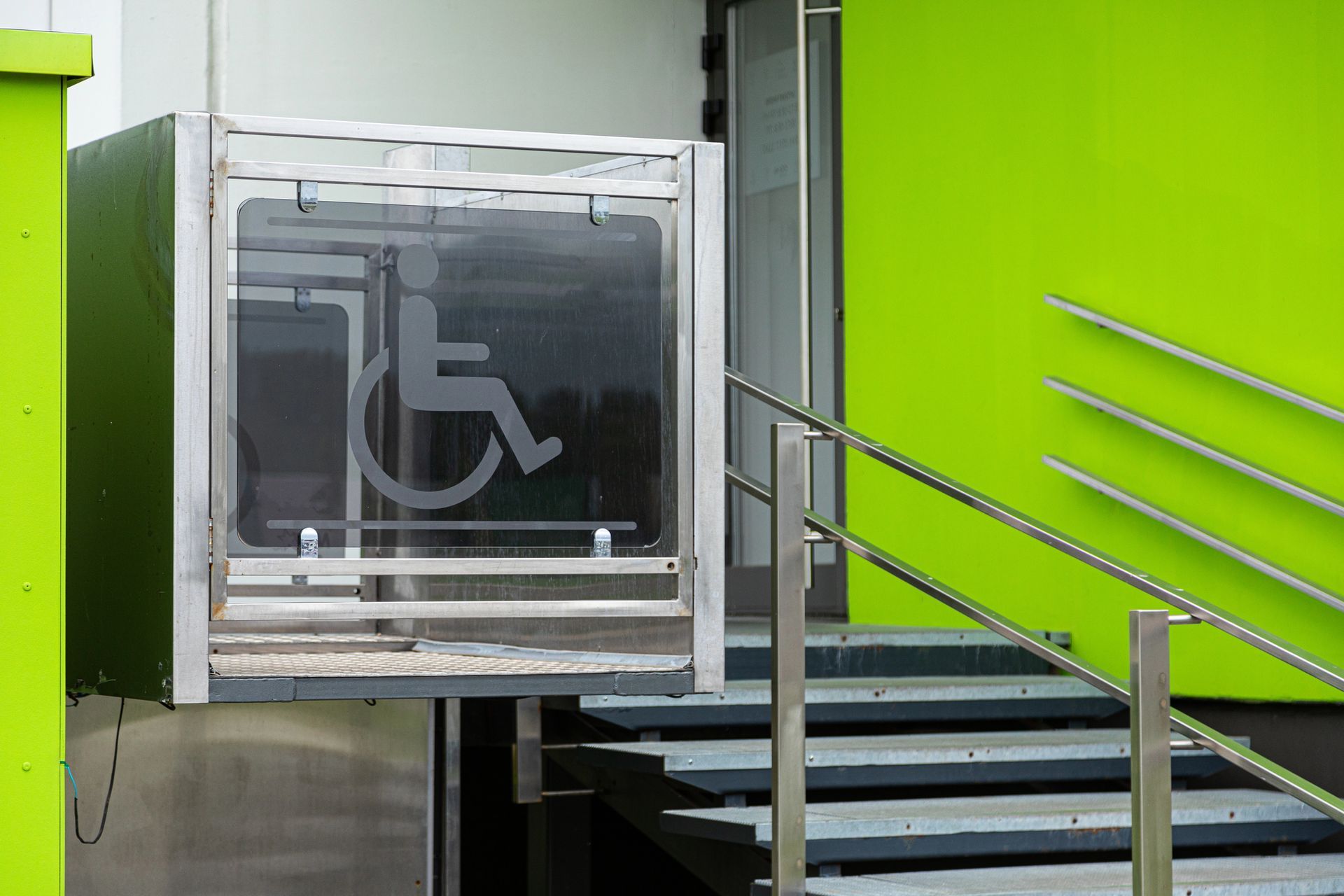 Un elevador de sillas de ruedas para accesibilidad ubicado junto a un conjunto de escaleras en la entrada de un edificio con paredes de color verde brillante.