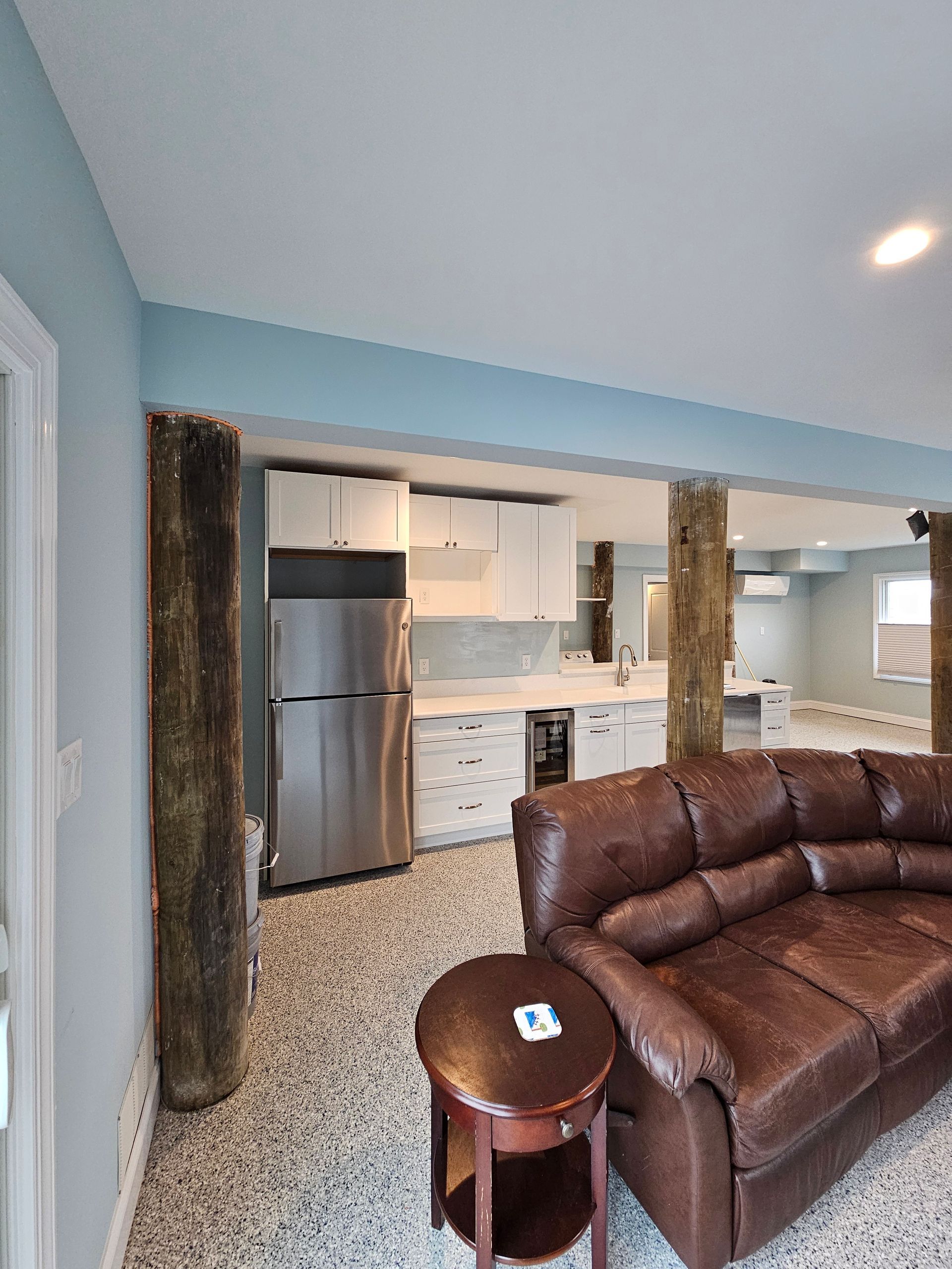 Living area with brown leather sofa, kitchen with white cabinets, and a tall, brown tree trunk column.
