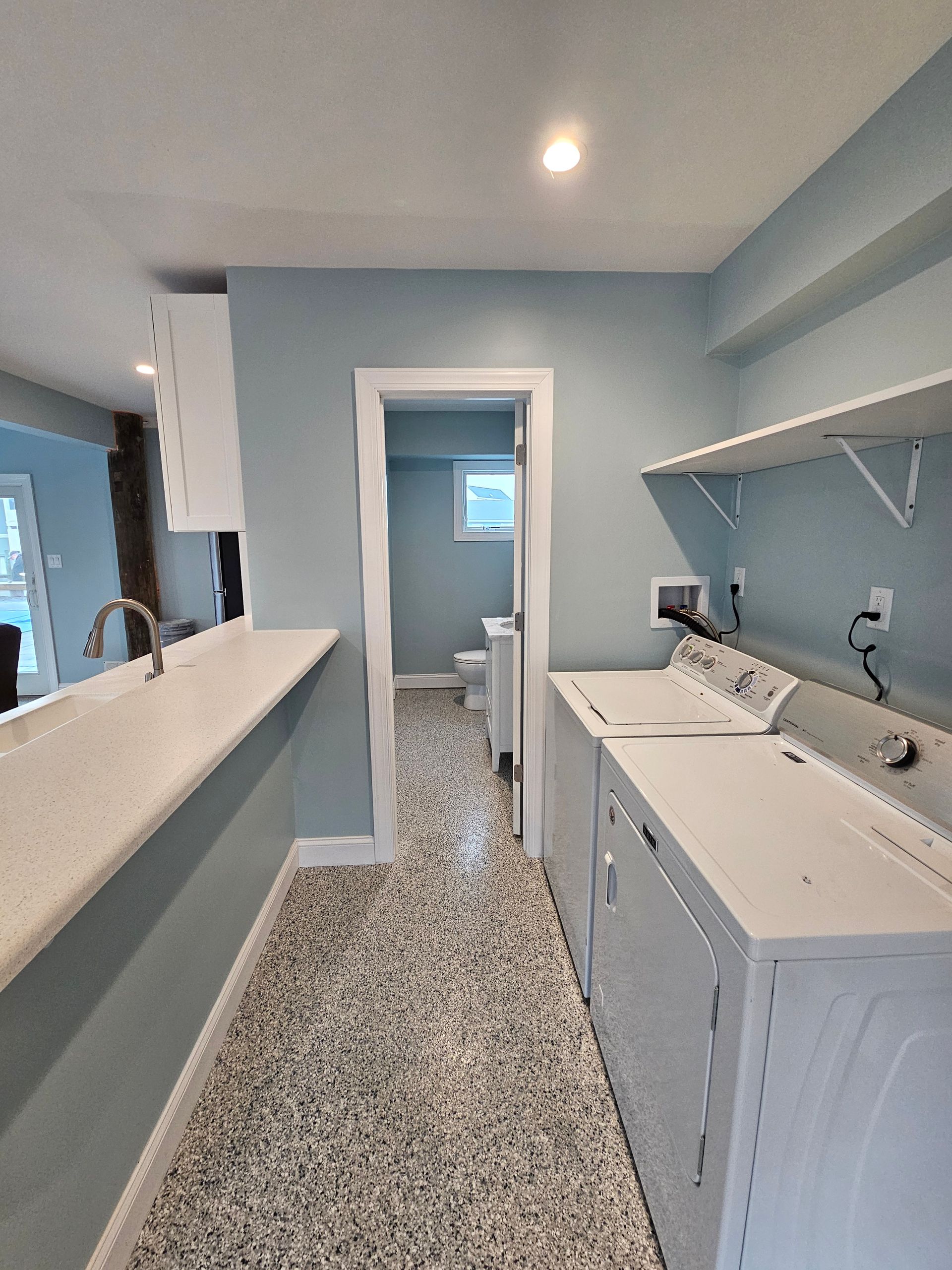 Laundry room with light blue walls, speckled floor, white appliances, and a hallway to a bathroom.