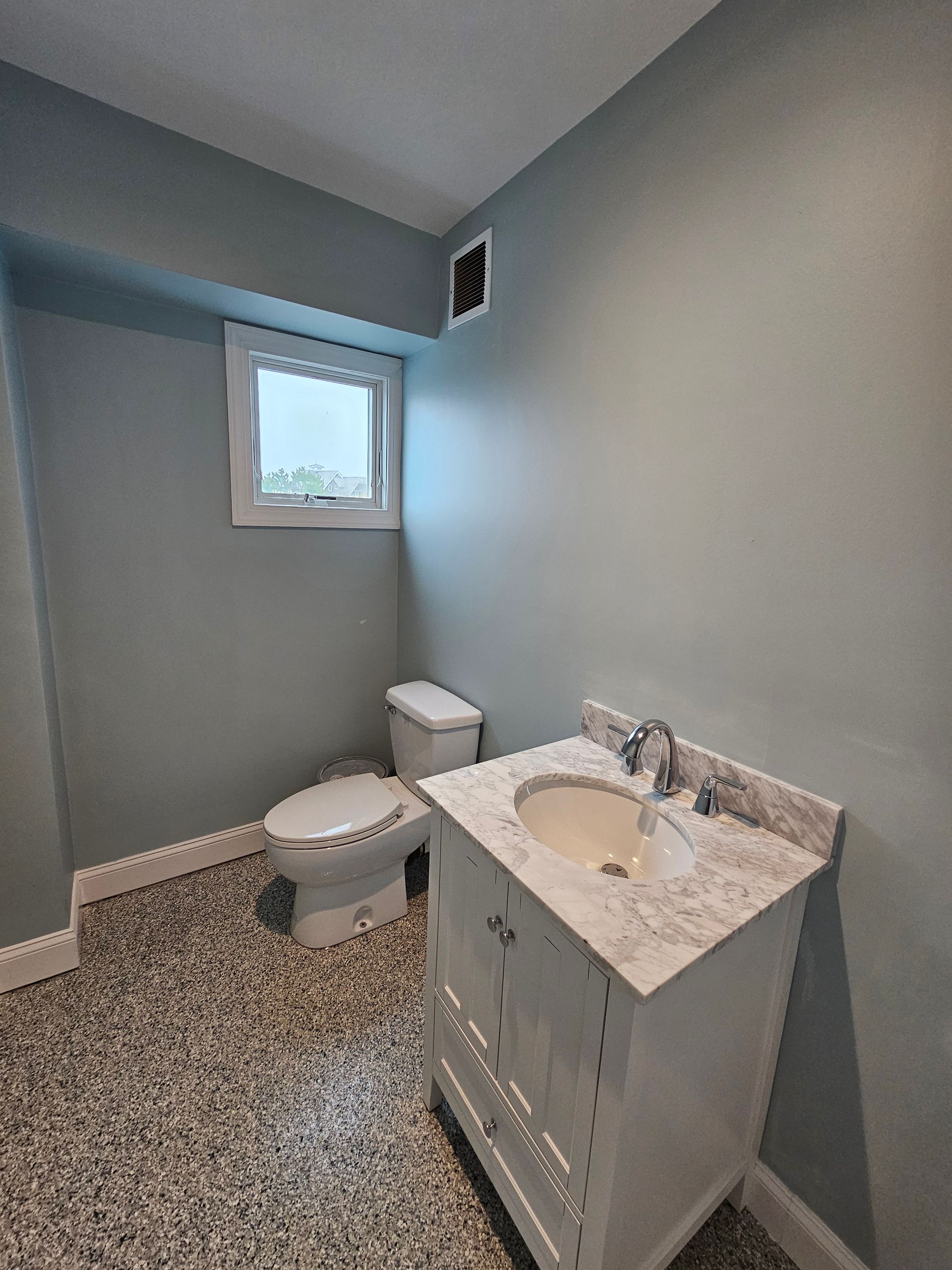Bathroom with toilet, vanity, window, and patterned floor. Light blue walls, white cabinet.