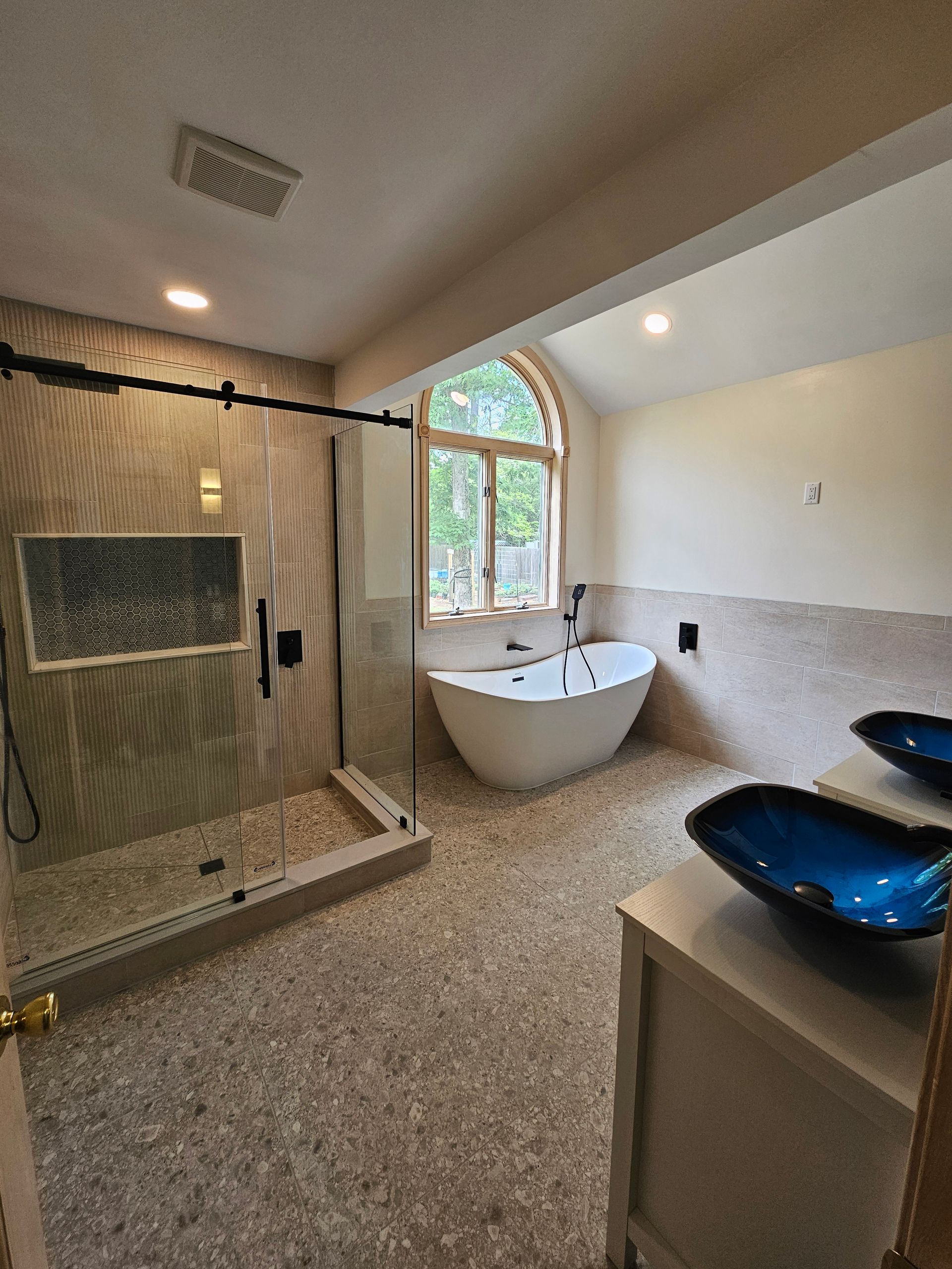 Modern bathroom with shower, bathtub, and blue vessel sinks. Pebble tile floor and arched window.