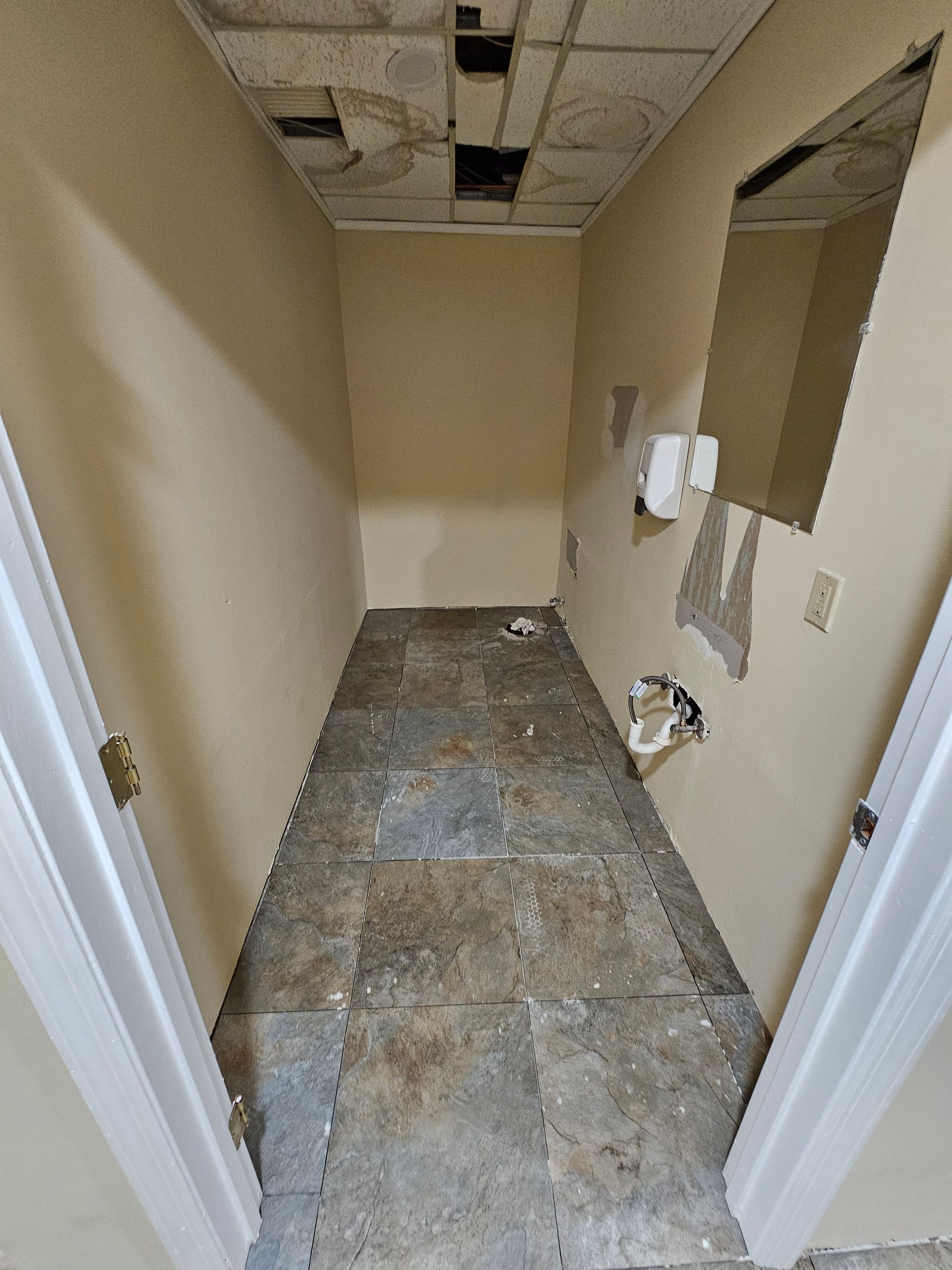 Narrow room with damaged floor and ceiling, plumbing fixtures, and two doorways. Beige walls and white door frames.