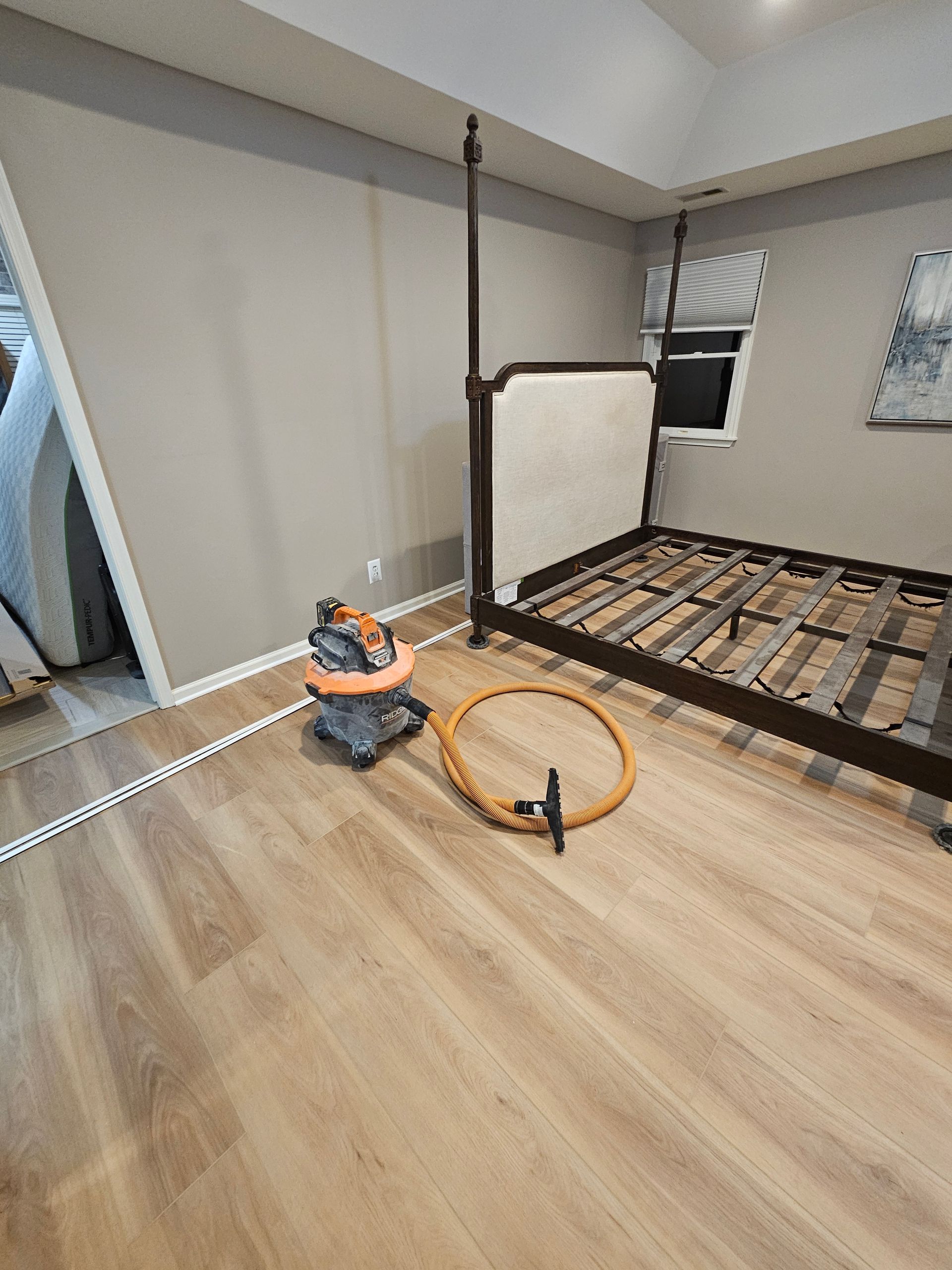 Bedroom with bed frame, vacuum, and hose on wood-look floor. Gray walls, window, and doorway visible.
