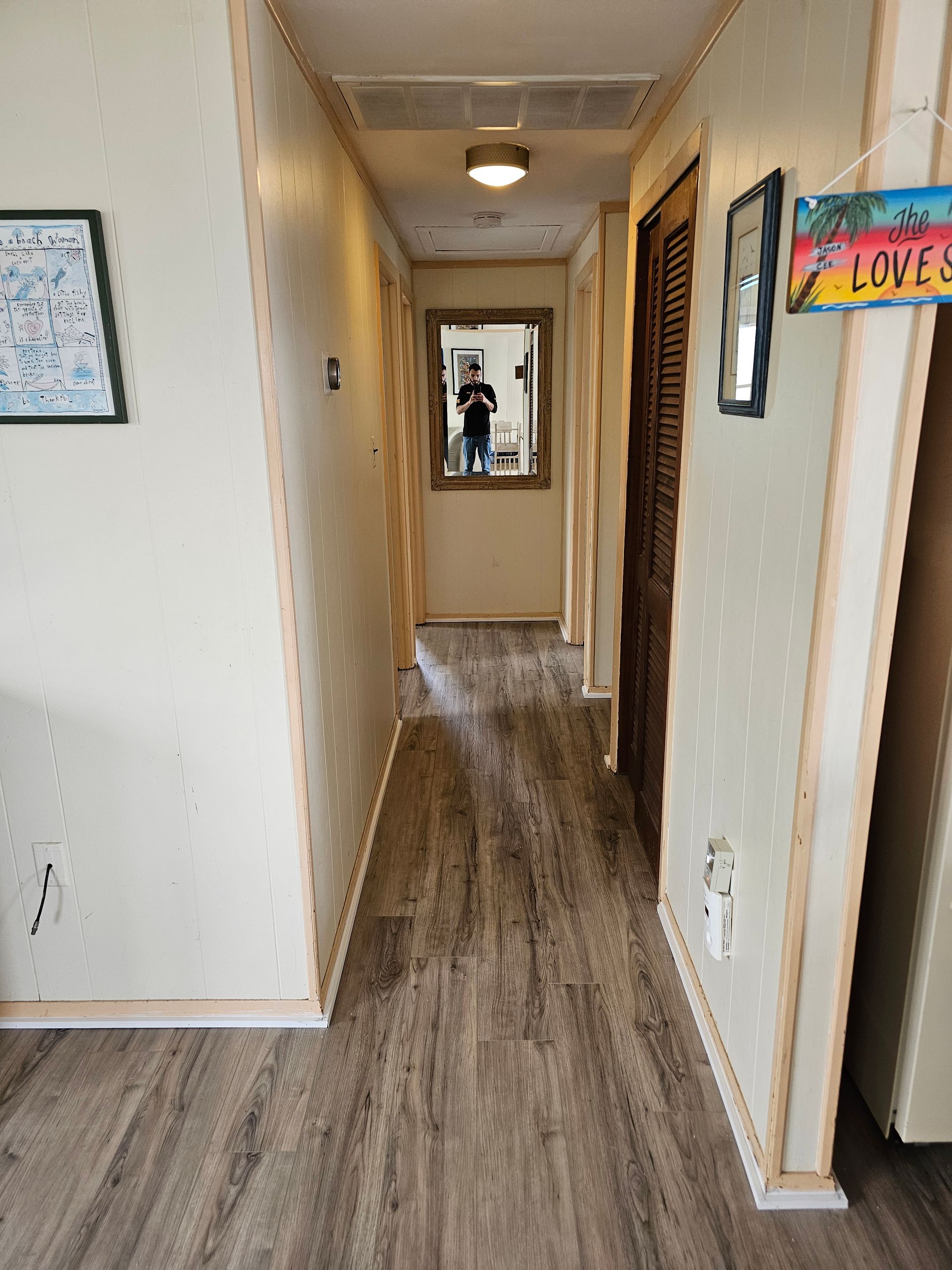 Narrow hallway with wood-look flooring, walls with trim, doors, and a mirror reflecting a person.