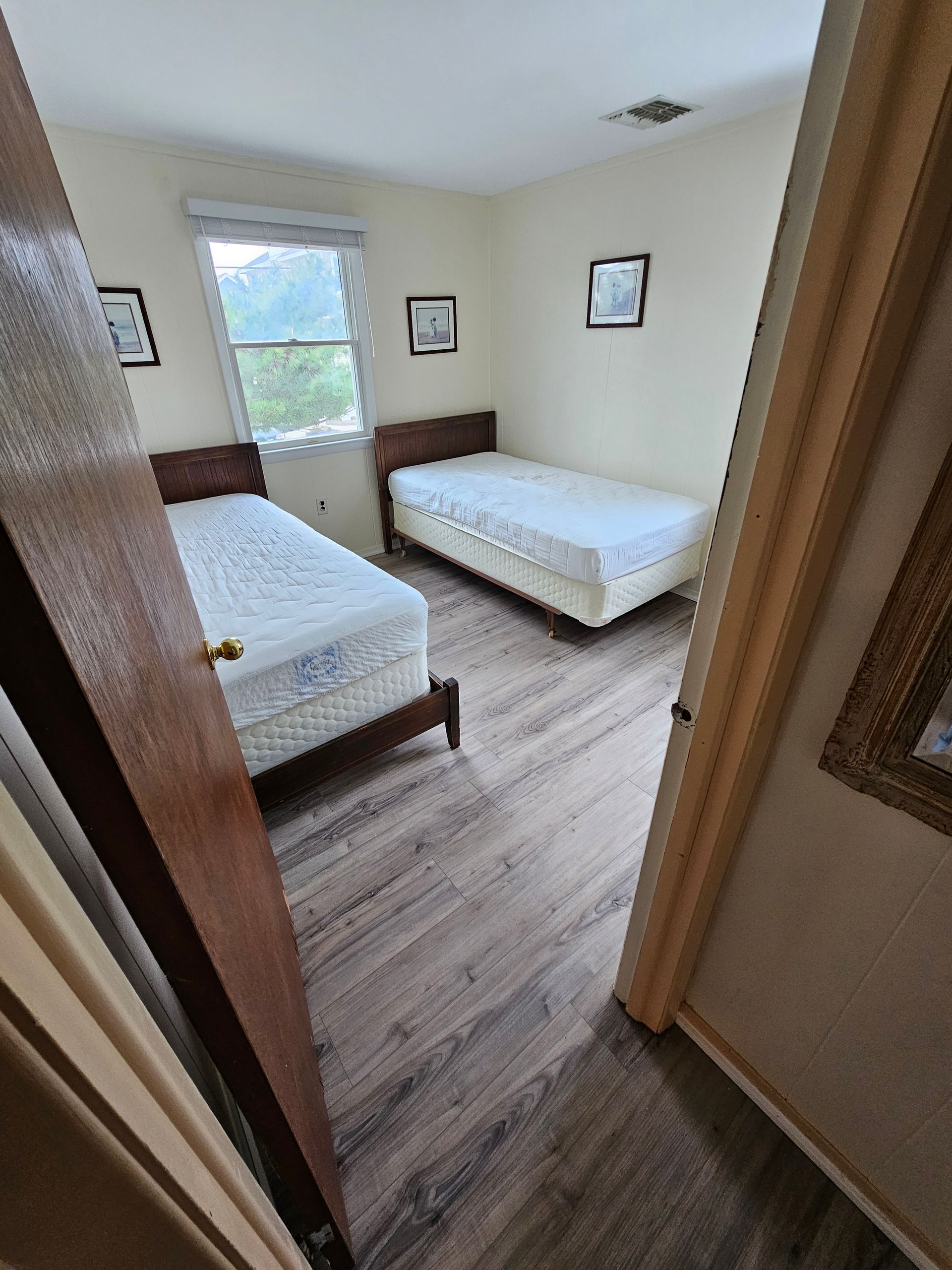 Two beds in a bedroom with wood floors, small window, and framed pictures on the walls.