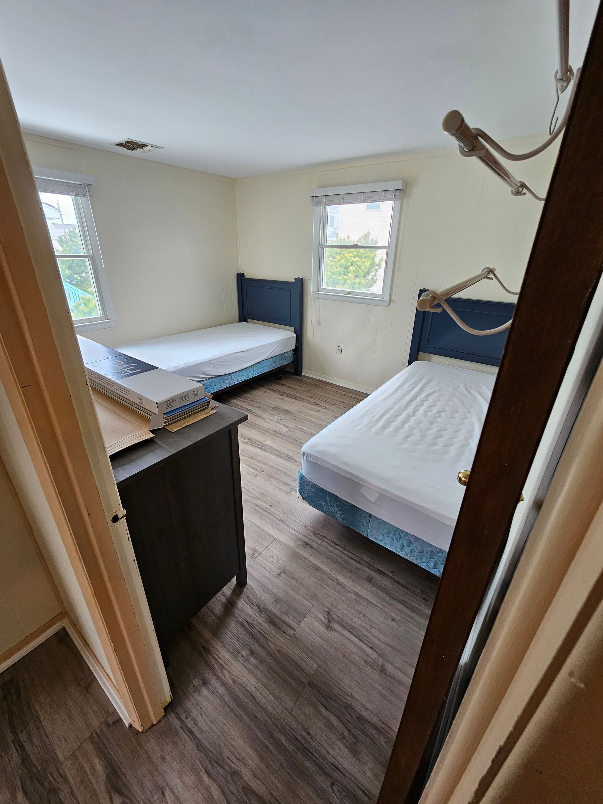 Bedroom with two beds, dresser, and two windows. Wooden floor, blue bedframes, white bedding.