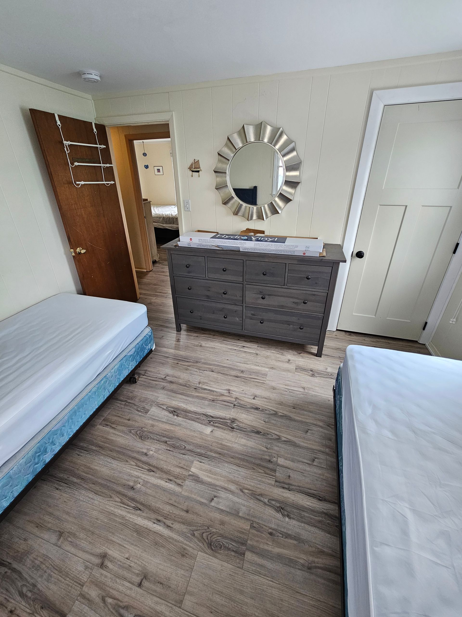 Bedroom with two beds, dresser, round mirror, and open doors. Light wood-look floor.