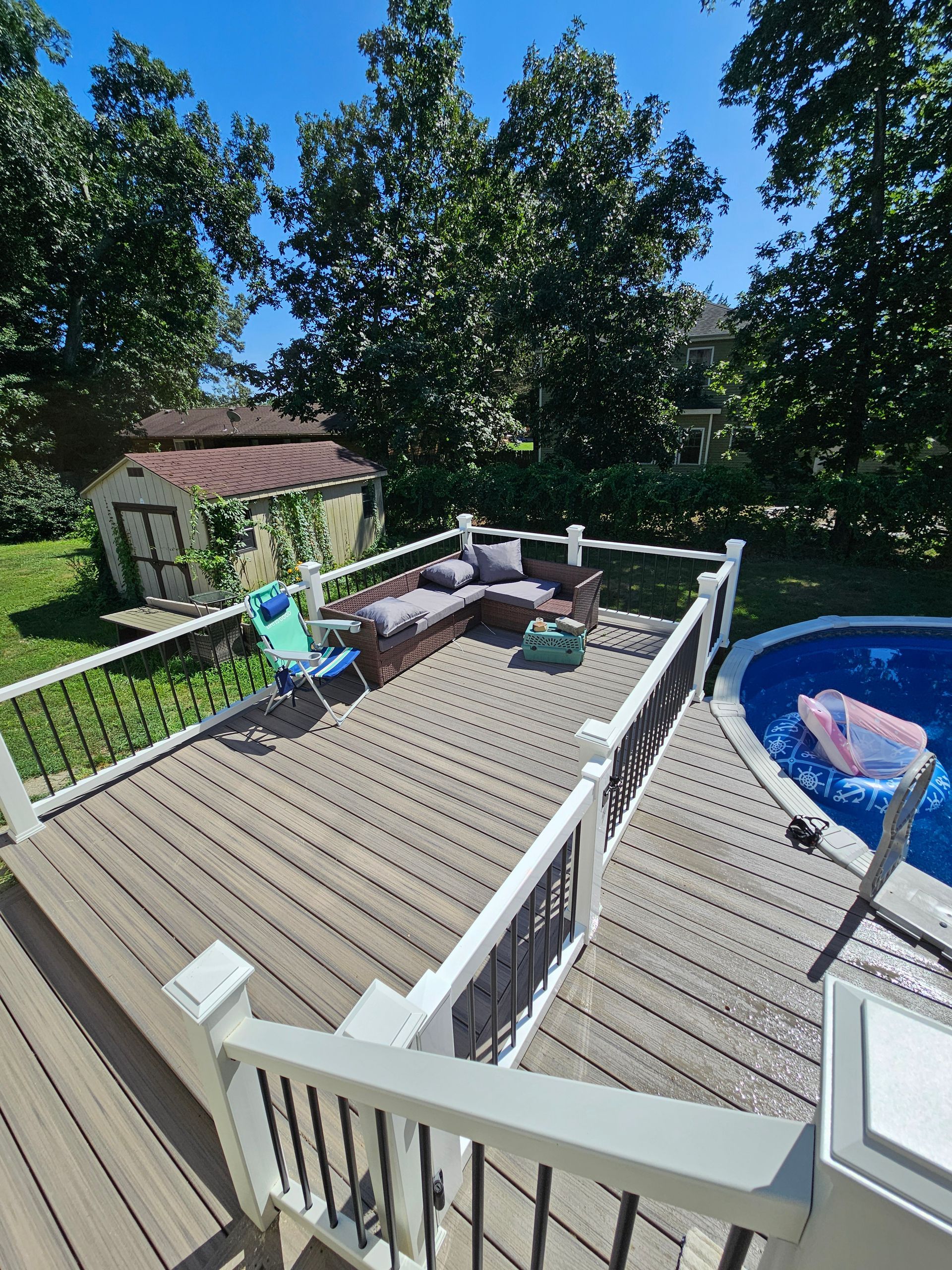 A backyard deck with seating, next to a pool. A small shed is visible in the background. Sunny day.
