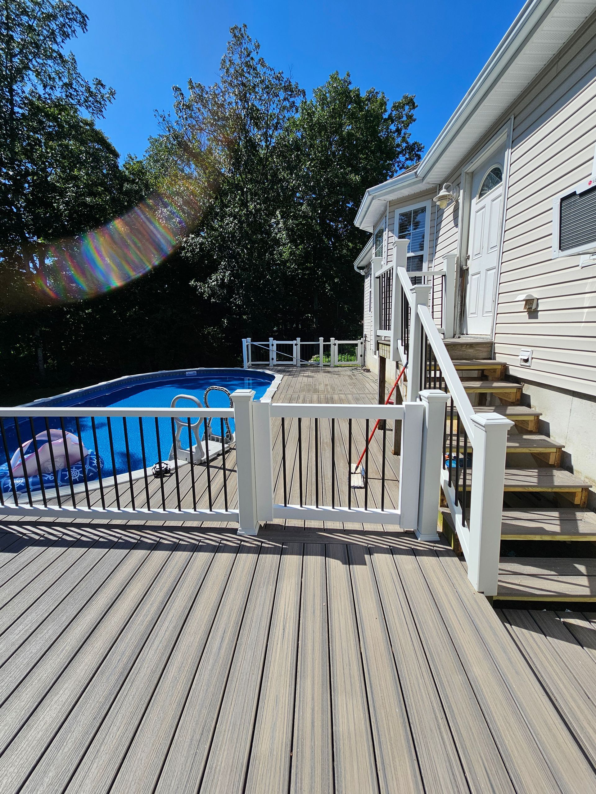 A deck with stairs leading to a house. A pool is behind a white picket fence on the left side of the house.