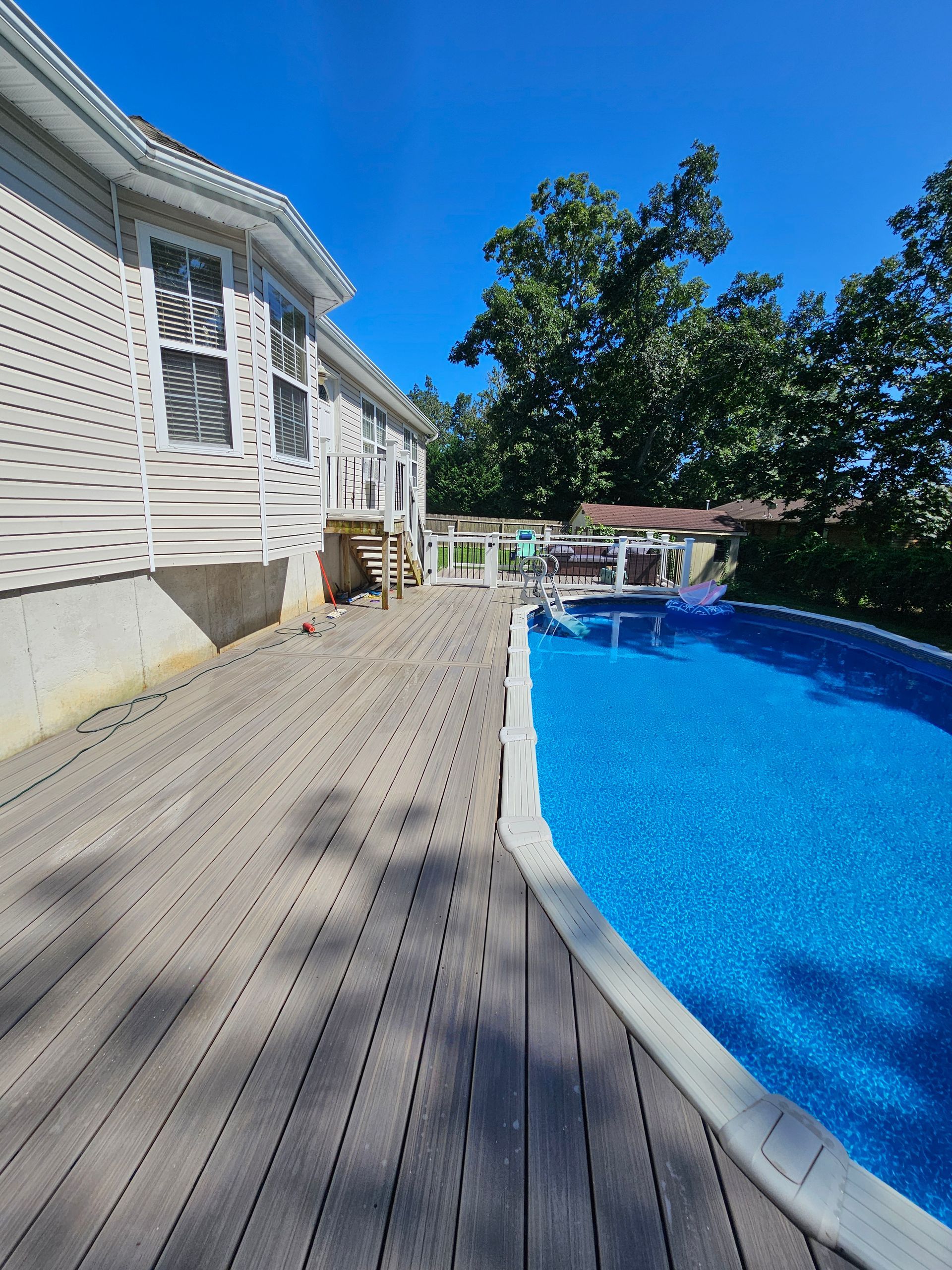 Wooden deck beside a blue pool next to a white house with blue sky.