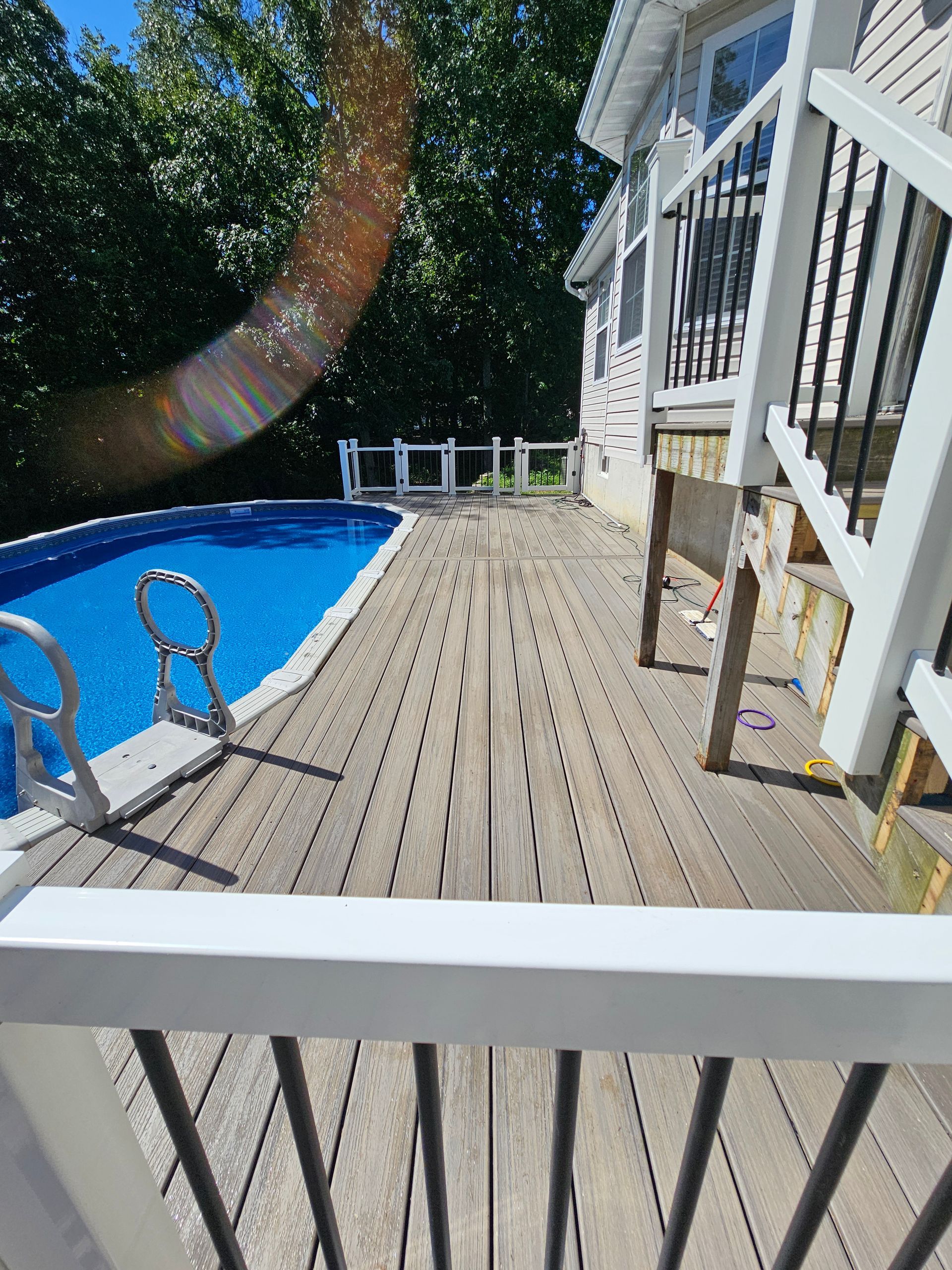 Wooden deck with a blue pool on the left and a white house on the right, sunny day.
