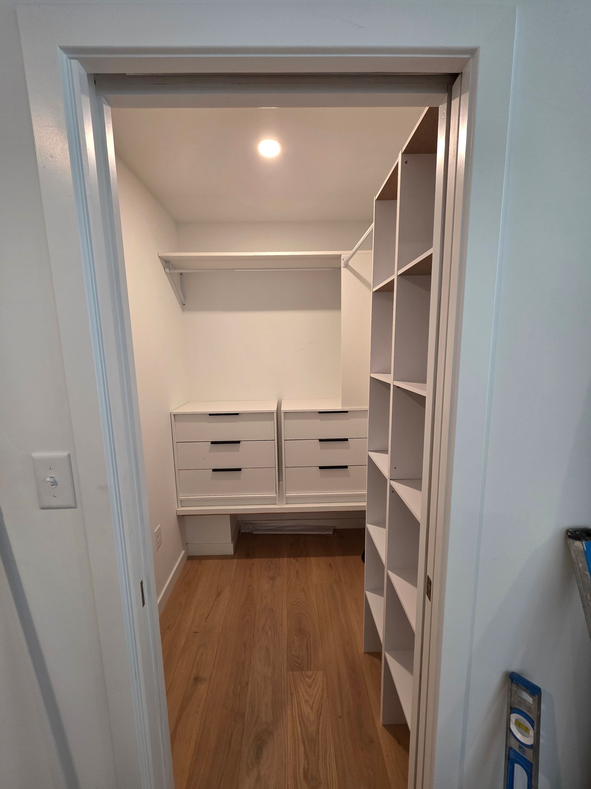 White walk-in closet with shelves, drawers, and wood flooring. Light fixtures visible.