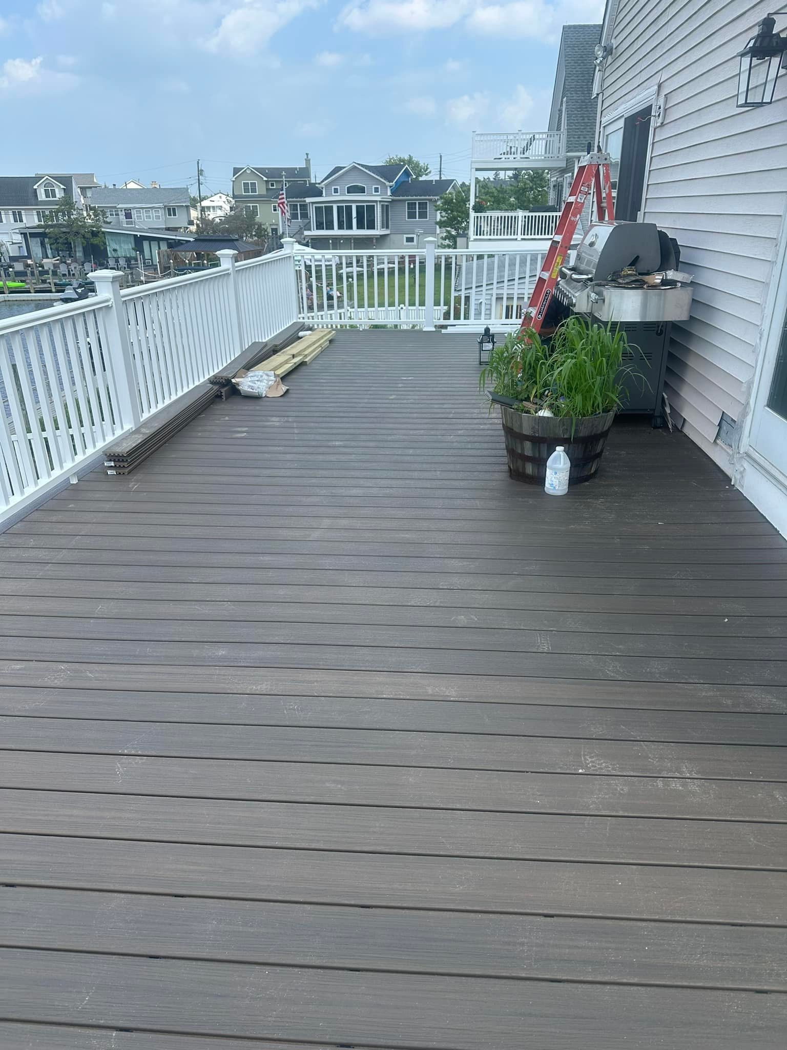 Wooden deck with white railing next to a house. A potted plant and grill are on the deck. Houses are in the background.