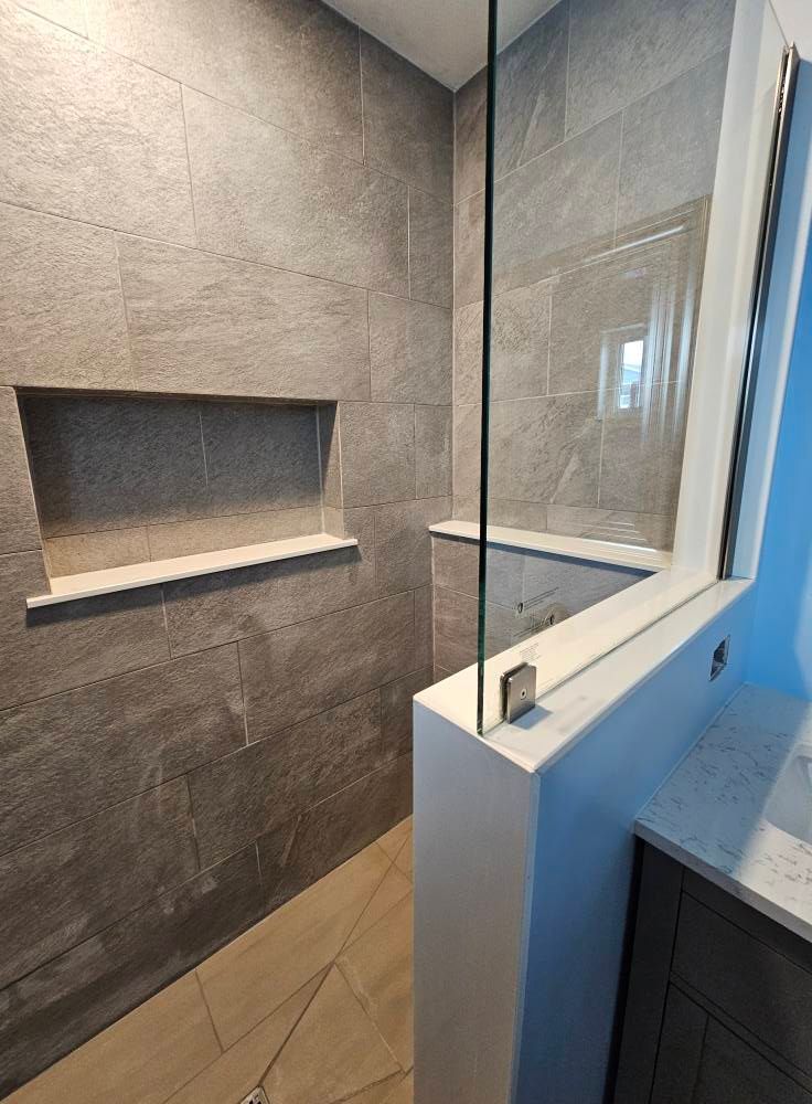 Shower with gray tiled walls, recessed shelf, glass partition, and white accents.