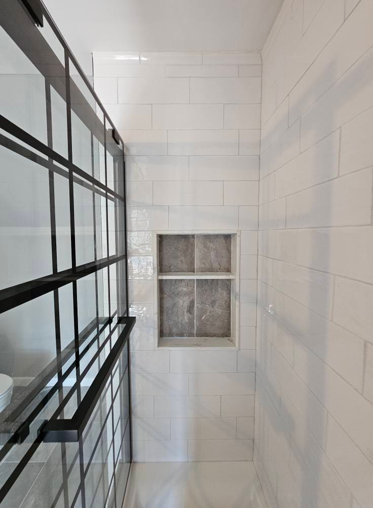 Bathroom with white tiled walls, glass shower door, and a built-in shelf. Black metal framing.