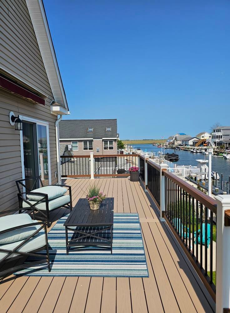 A sunny deck overlooking a waterway, with seating, rug, and view of houses and boats.
