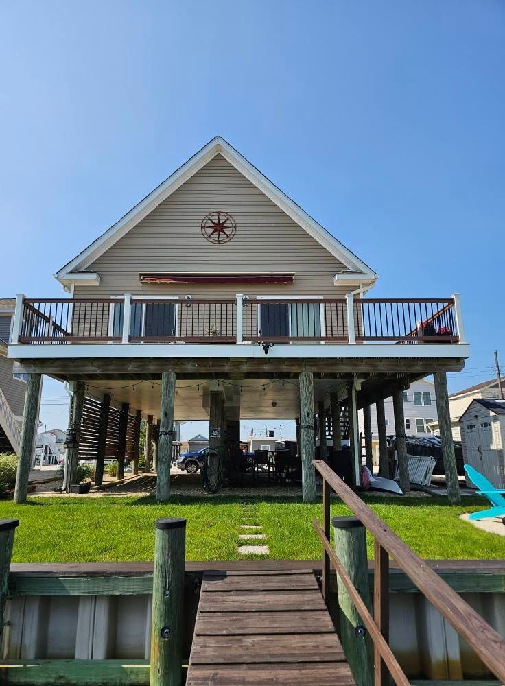 Beach house on stilts with a wooden deck, small lawn, and dock on a sunny day.