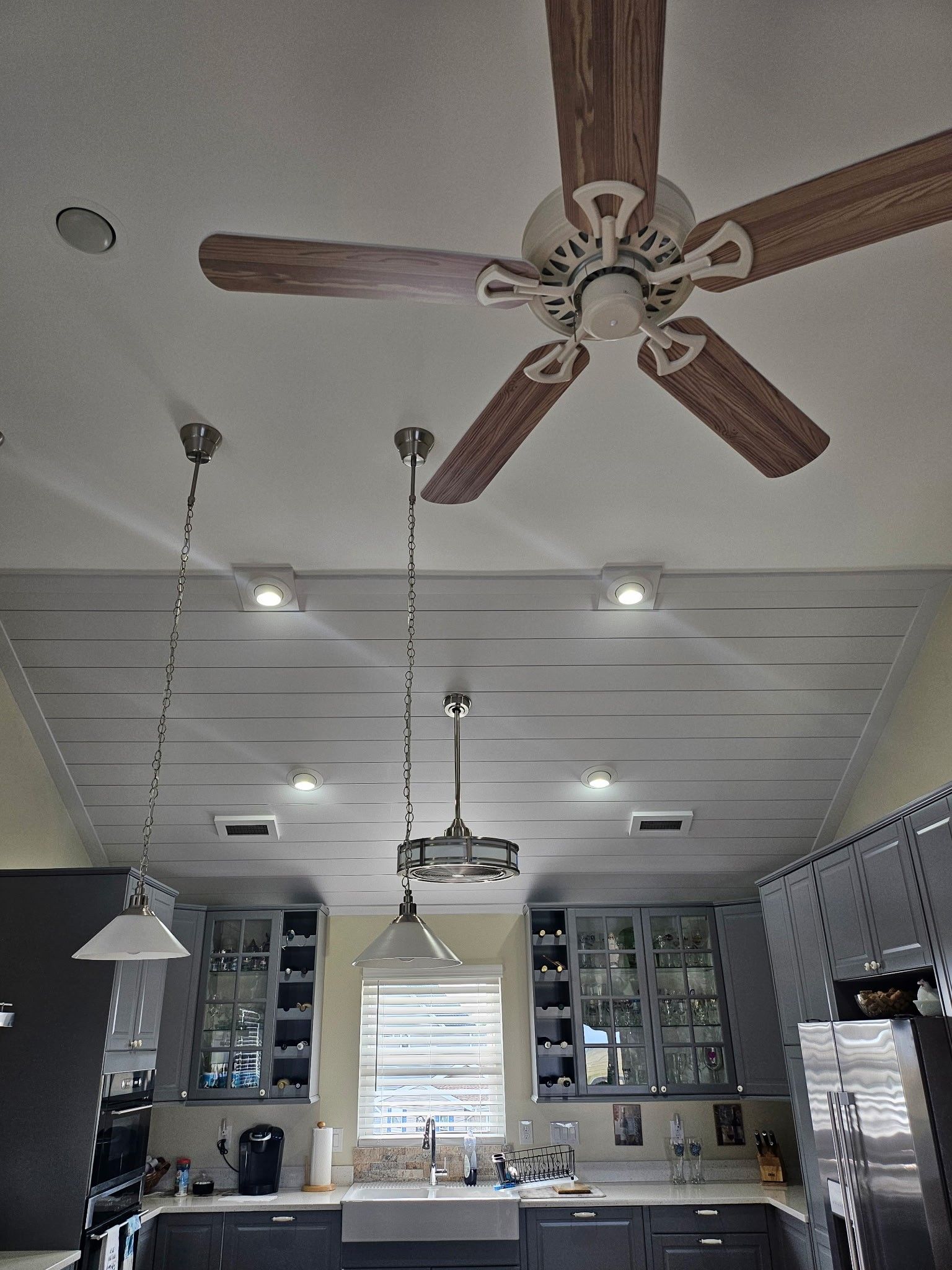 Kitchen ceiling with a ceiling fan and pendant lights over a countertop with cabinets and a window.