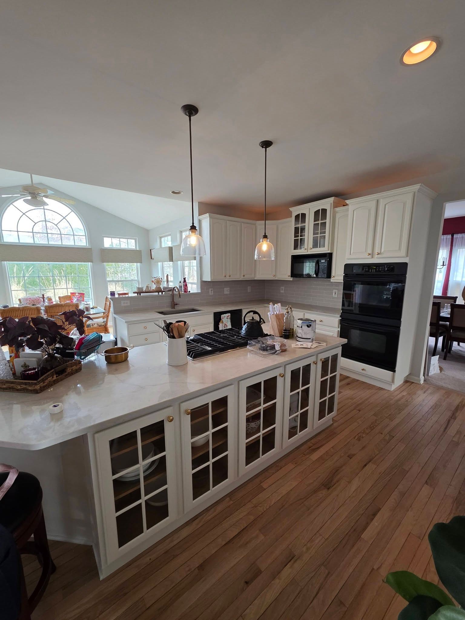 Kitchen with light wood floors, white cabinets, and island with glass-paneled doors; pendant lights hang above.