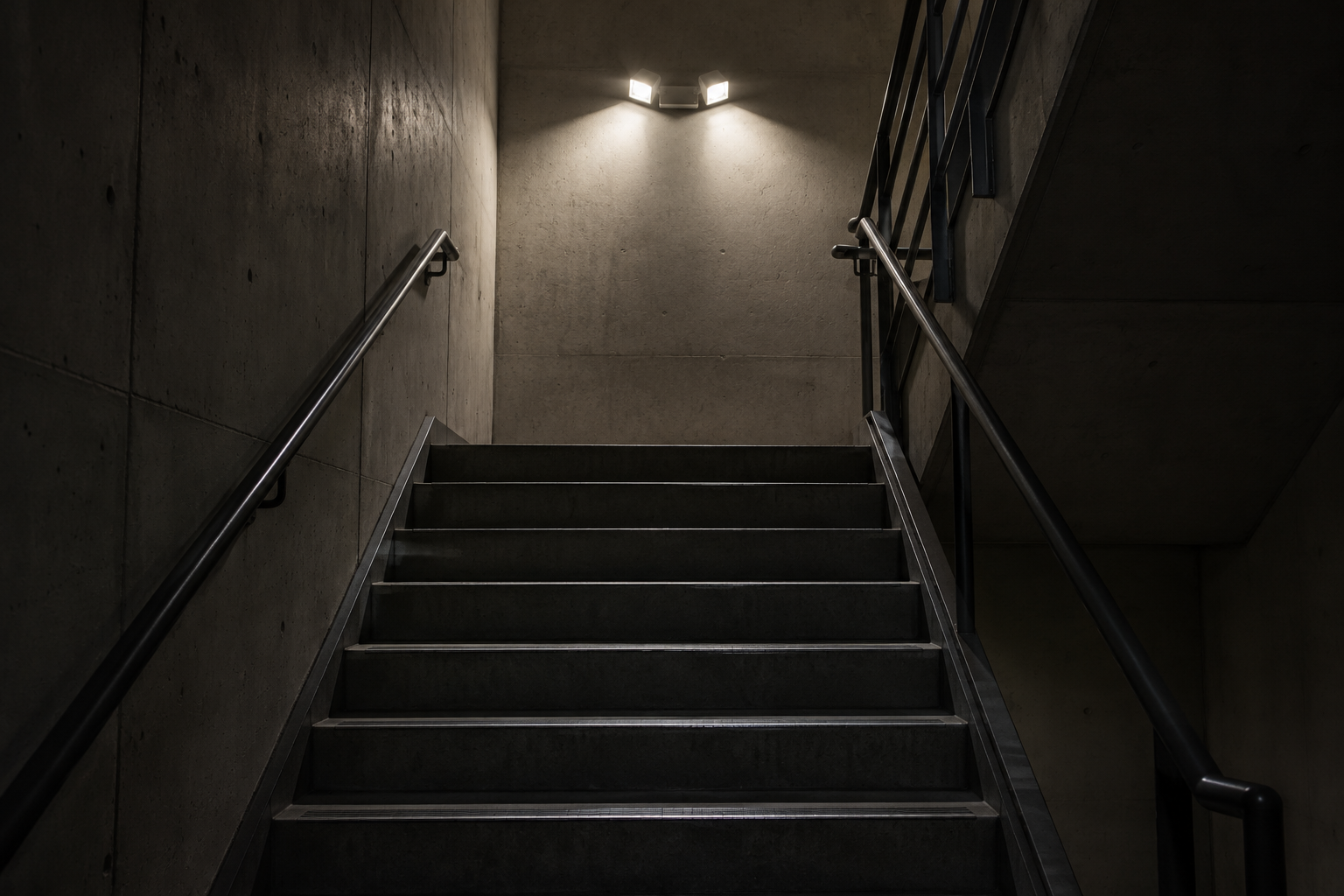 Dim stairwell with metal railings and wall lights leading upward in a dark concrete corridor