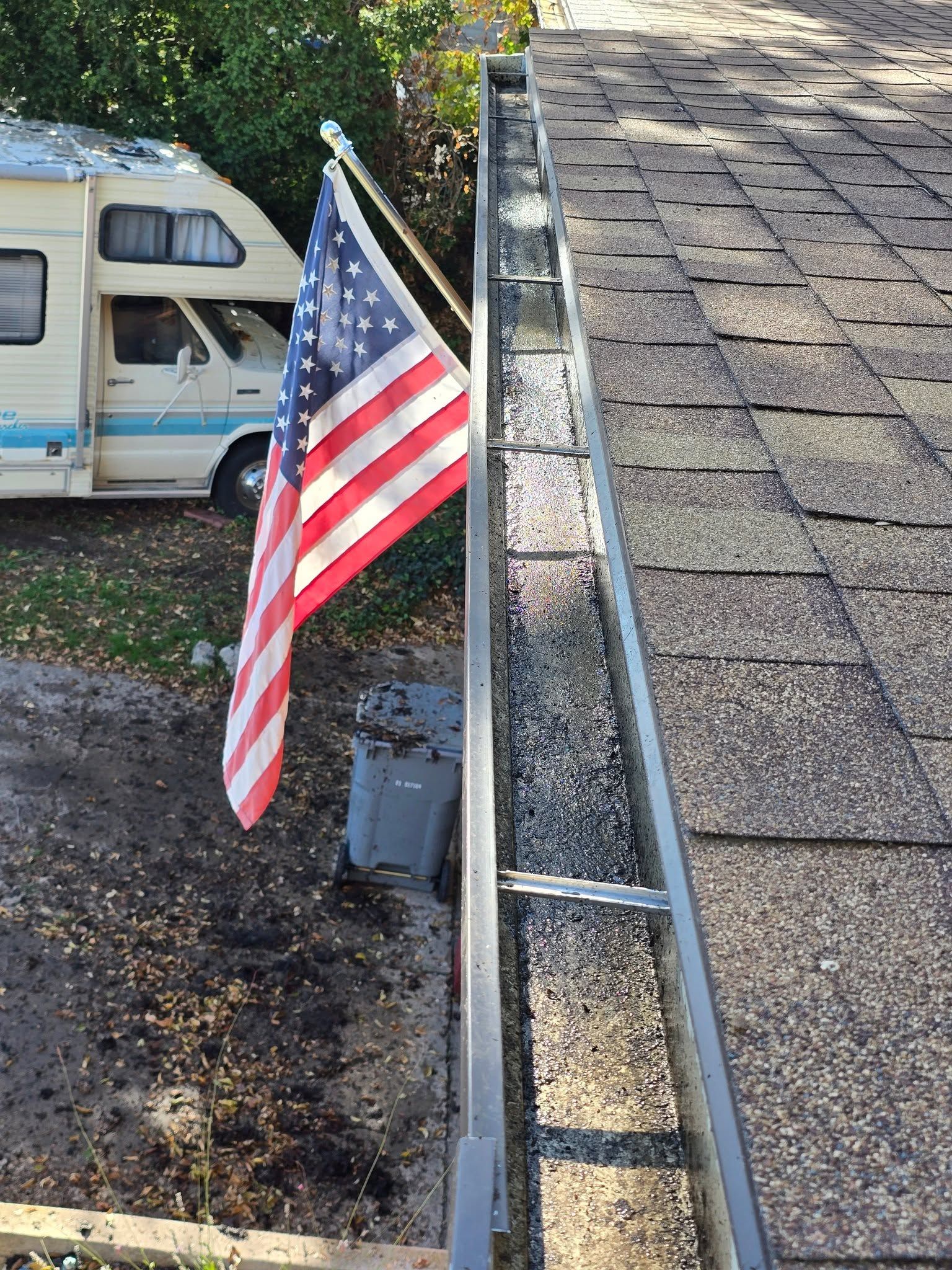 American flag mounted beside a roof gutter above a parked camper van and gravel driveway