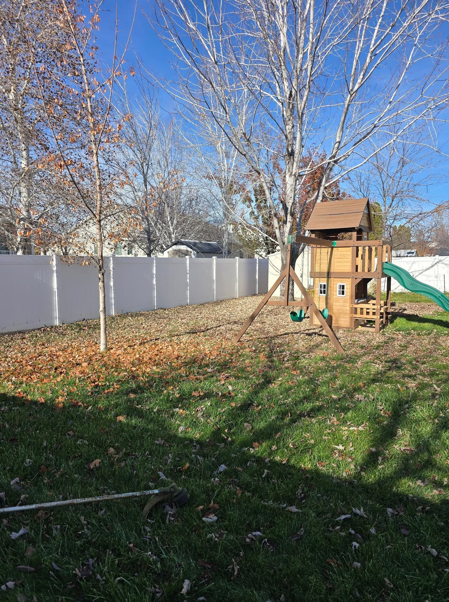 Backyard with white fence, leafless trees, green grass, and a small wooden playhouse/play set.