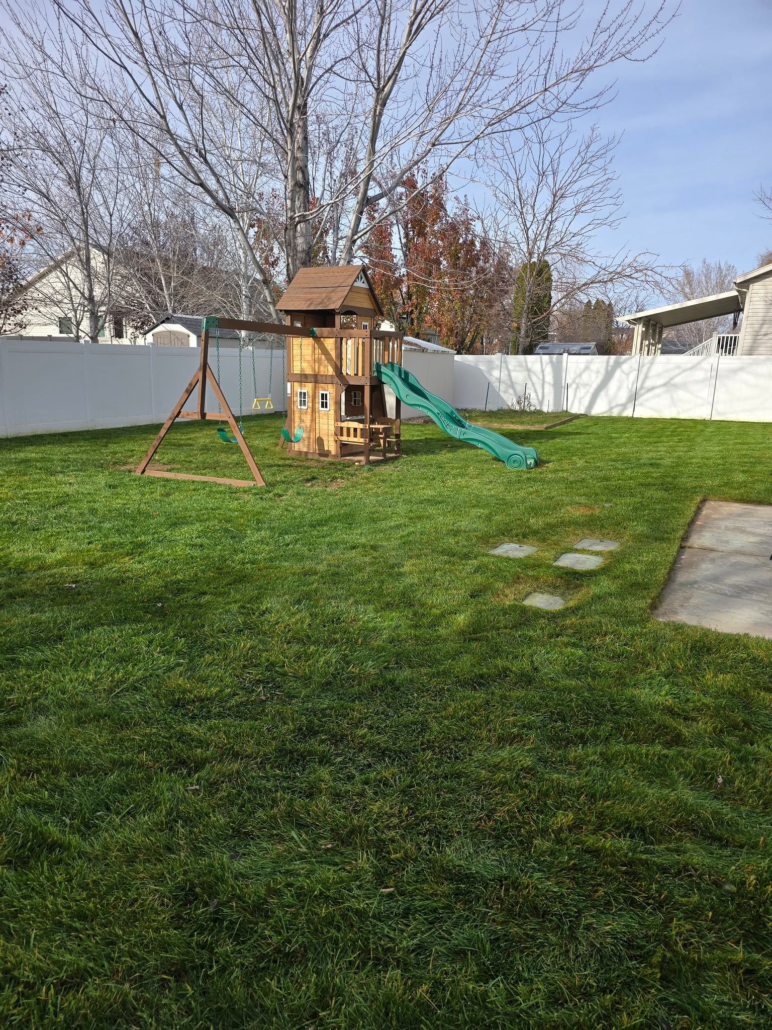 Backyard with a wooden playset, green slide, tree swing, and white fence under a bare tree.