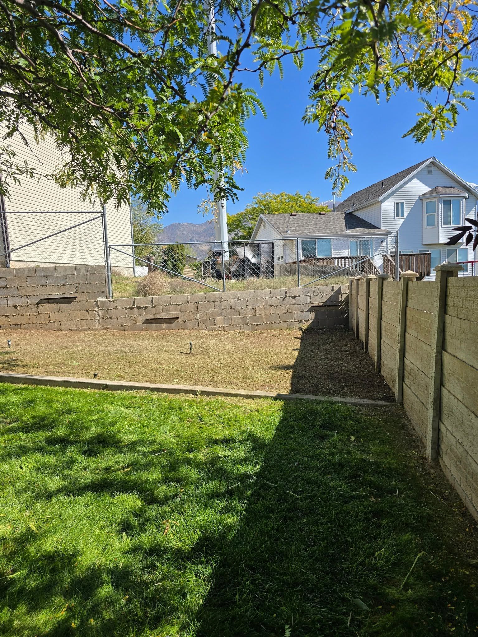 Backyard with grass, retaining wall, wooden fence, and two houses under a bright blue sky