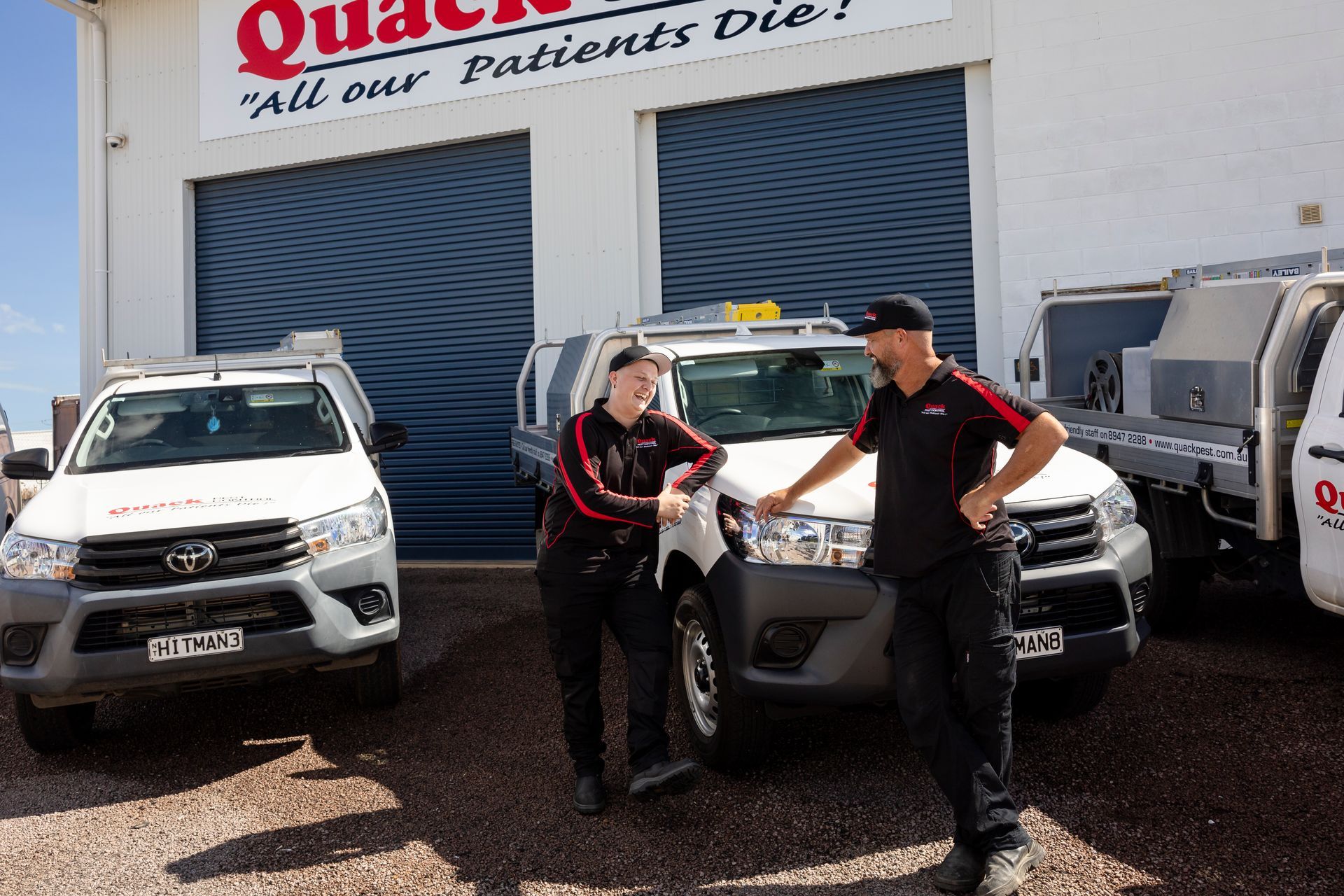 Two People in Work Attire Stand Beside White Service Trucks in Front of a Building — Quack Pest Control in Stuart Park, NT