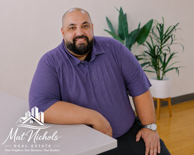 Man in a purple polo smiling beside a white table, with potted plants in a bright office.