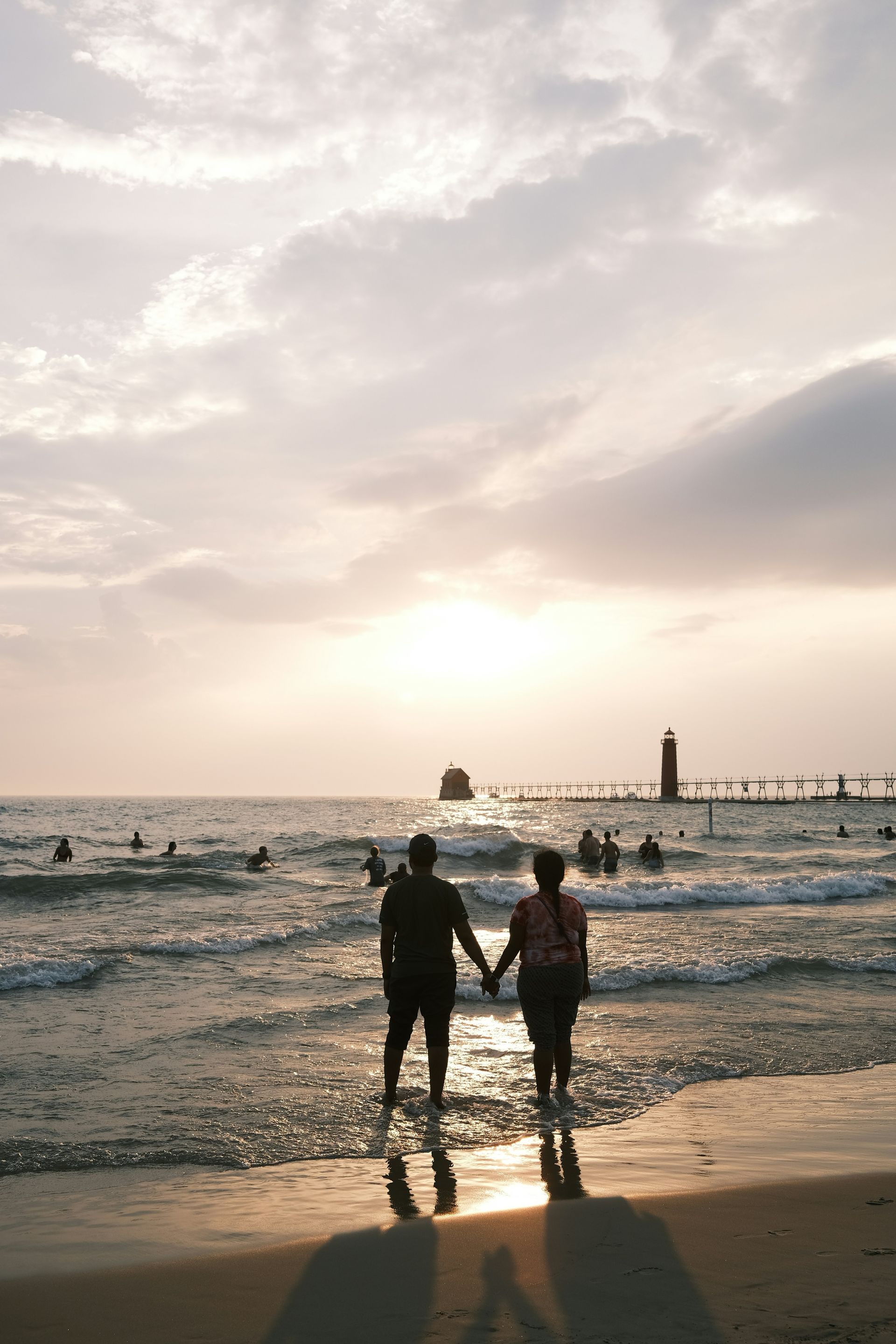 Couple holding hands at sunset on a beach, with waves and a lighthouse in the distance