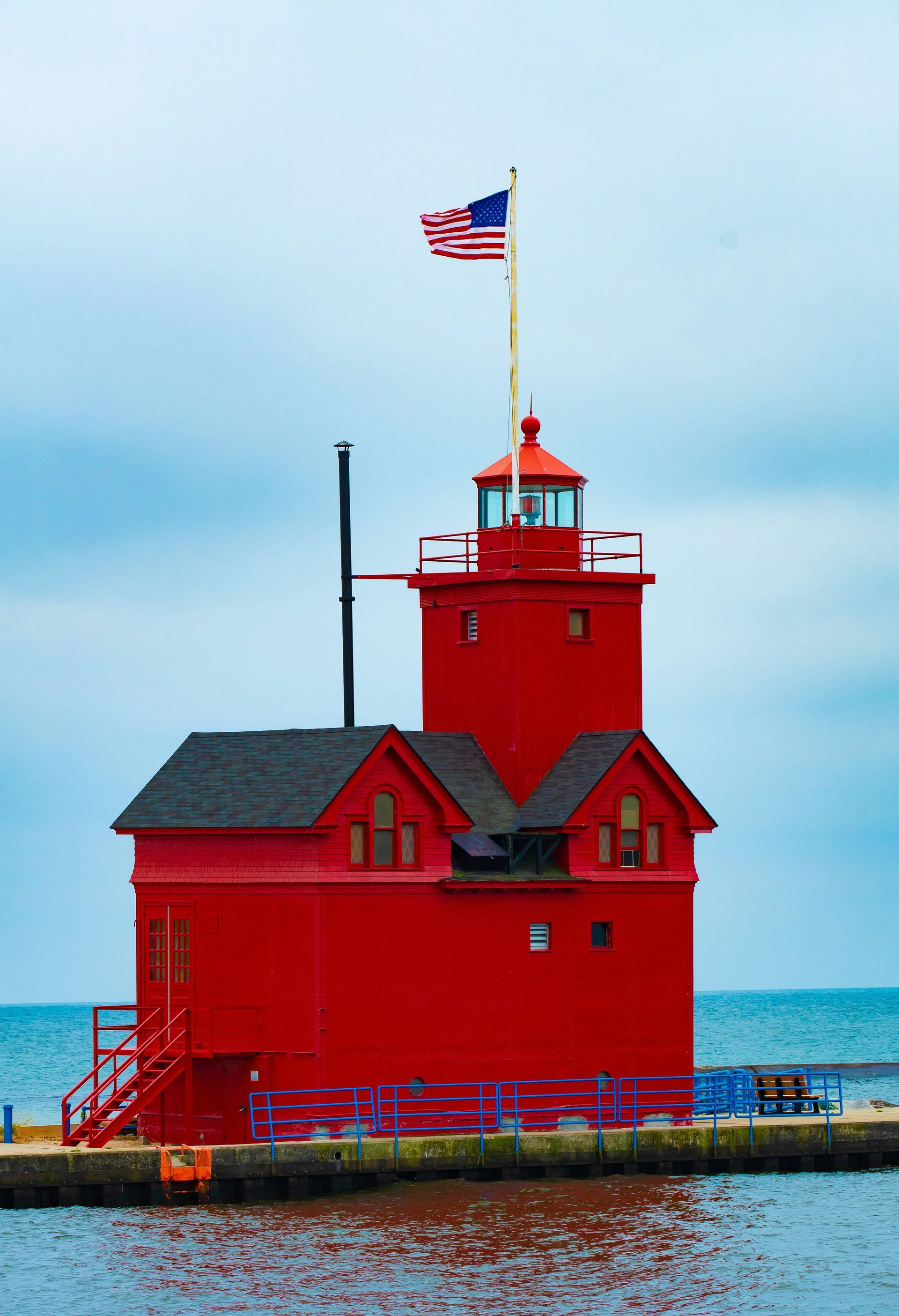 Bright red lighthouse with an American flag on a pier by the water under a cloudy sky