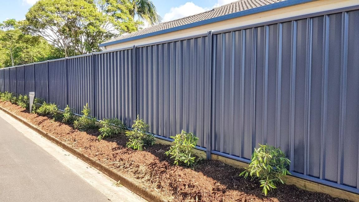 Blue metal fence along a road with small green bushes planted along the base.