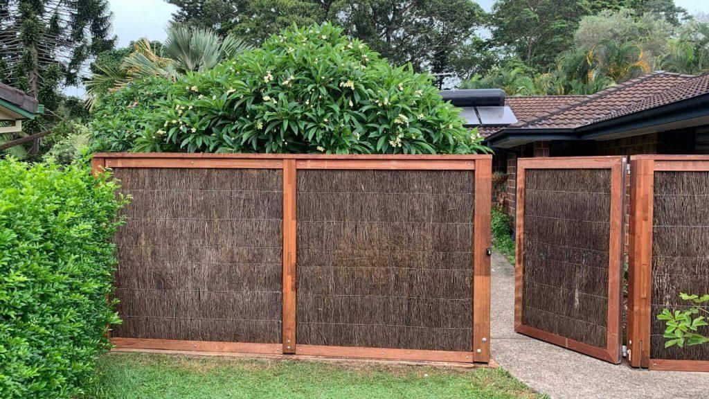 Brown reed fence with wooden frame in a yard, gate open, green foliage.