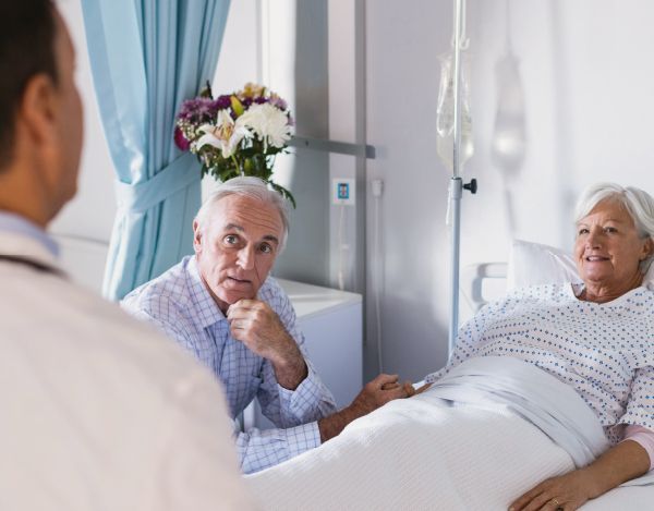 An elderly couple is laying in a hospital bed talking to a doctor.