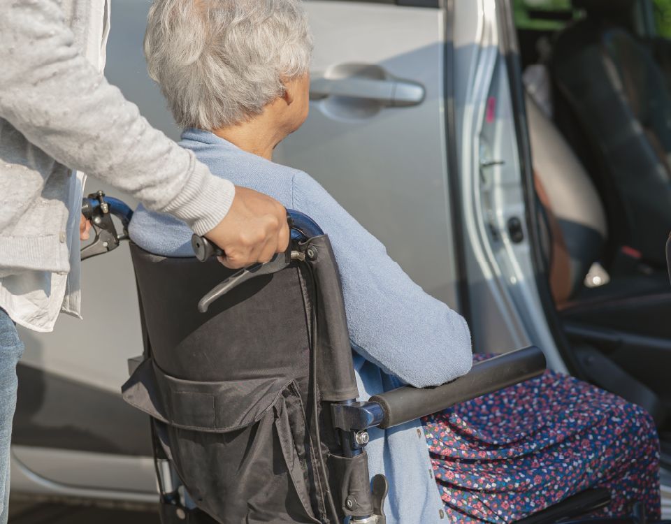 An elderly woman in a wheelchair is being pushed into a car.