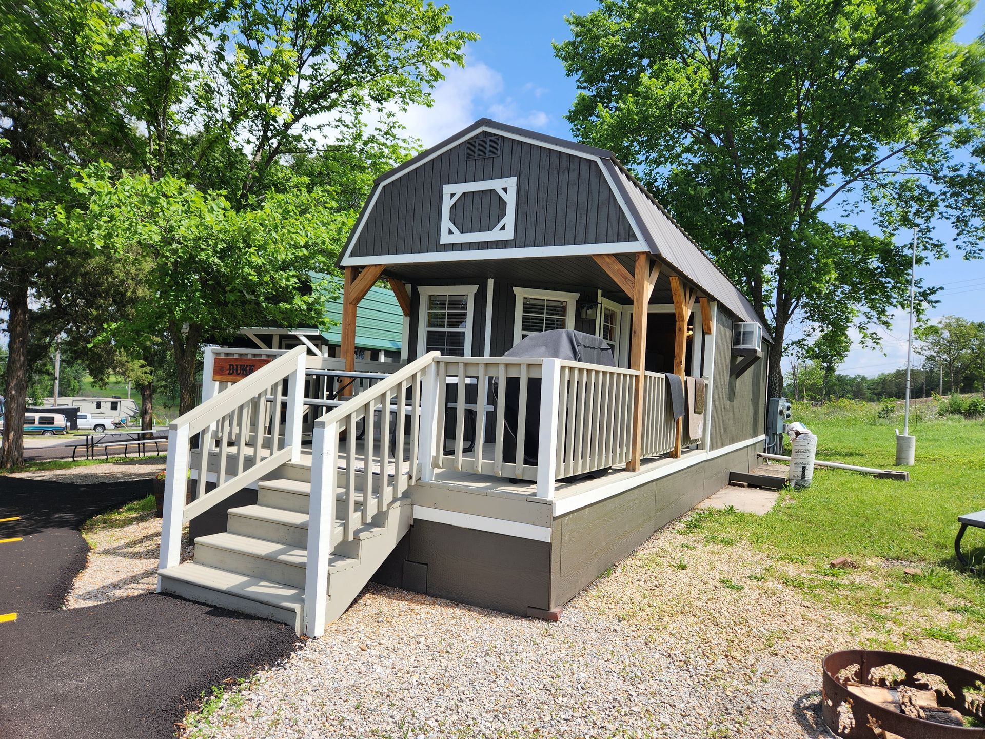 Small gray barn-style cabin with front porch and ramp, surrounded by trees and gravel