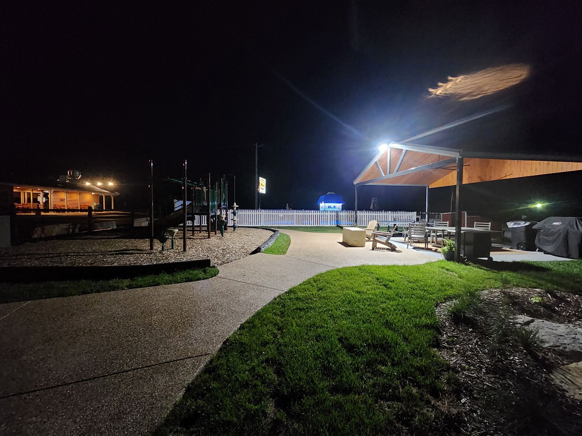 Nighttime outdoor plaza with lit walkways, grassy areas, and a shelter near a building.