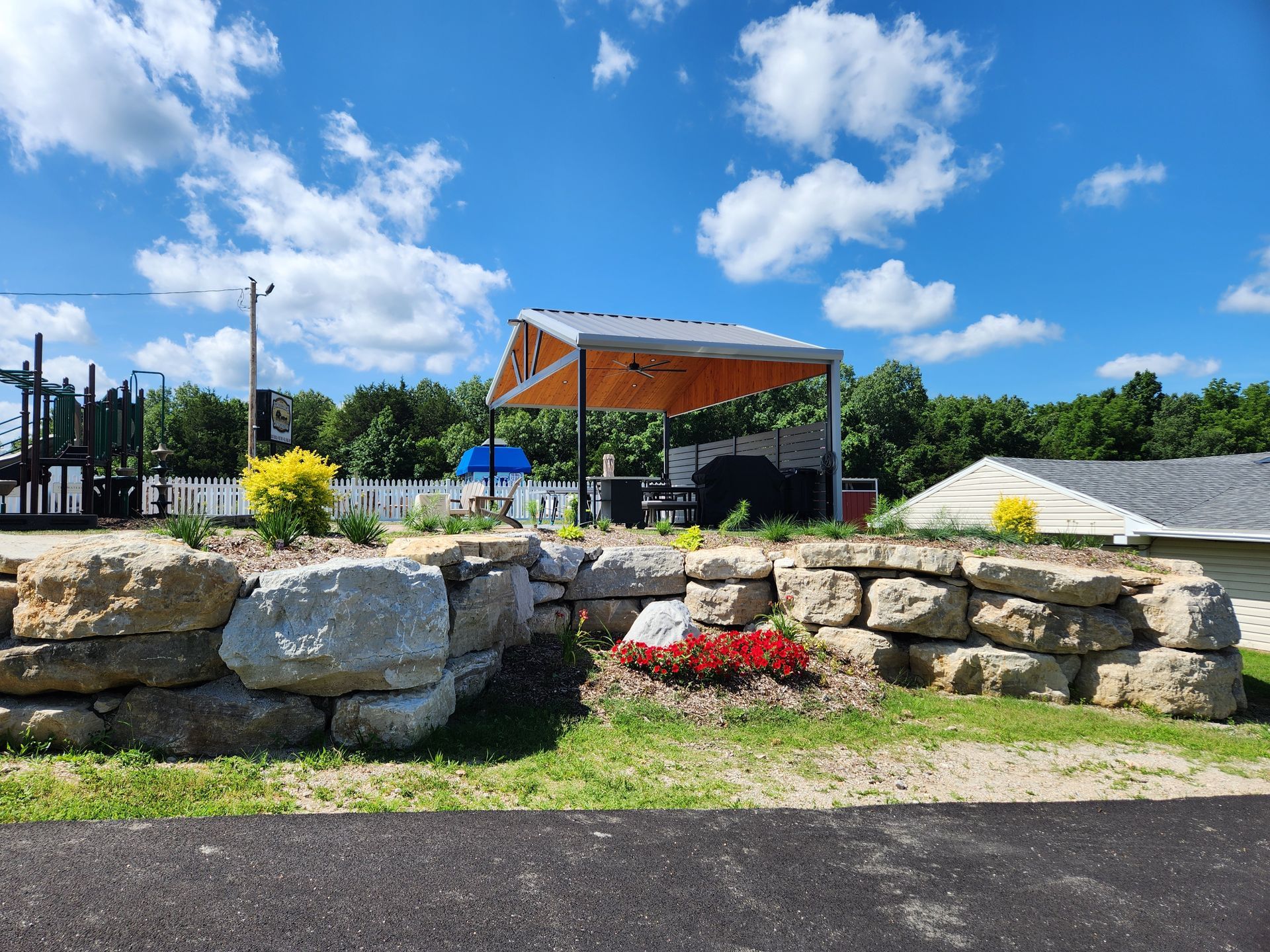 Stone garden wall with red flowers, wooden pavilion, and blue sky with clouds in a park