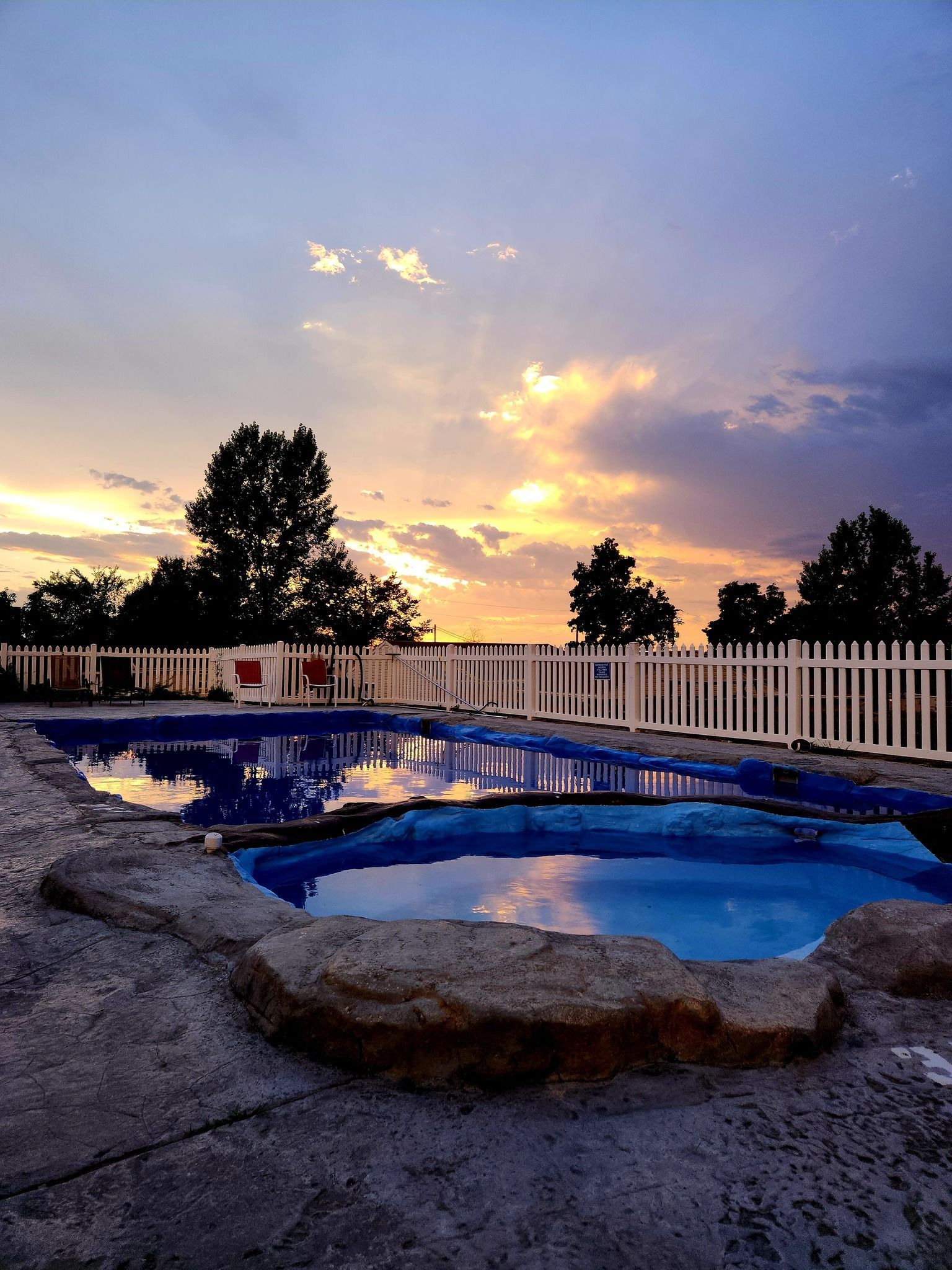 Backyard pool at sunset with glowing clouds, white fence, trees, and stone patio