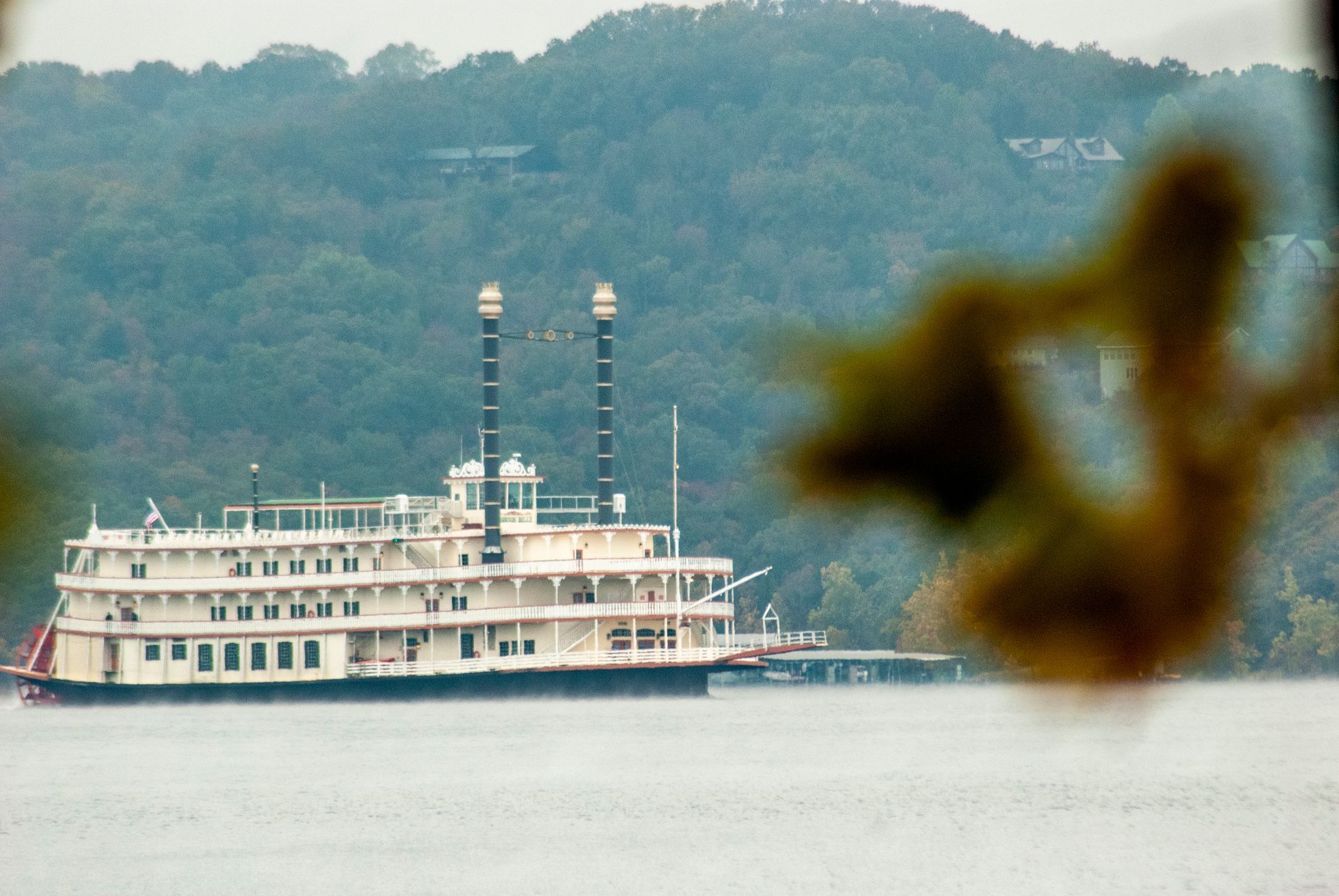 A white riverboat cruises on calm water with wooded hills in the background.
