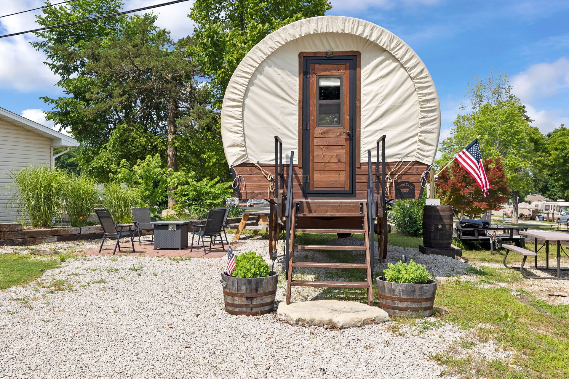 Tiny cabin with front steps, gravel yard, patio chairs, and an American flag under a blue sky.