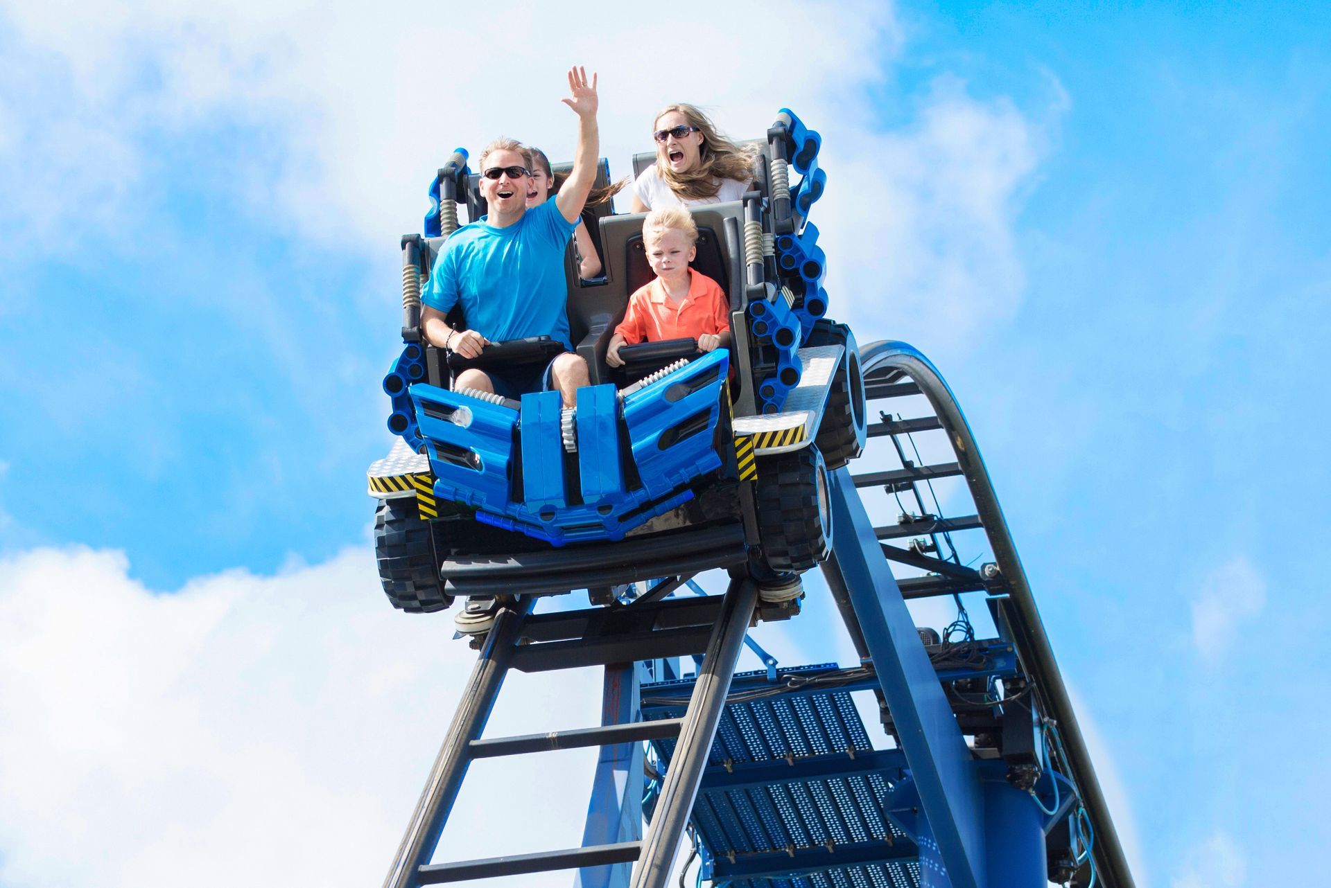 People riding a blue roller coaster, smiling and raising their hands against a bright sky