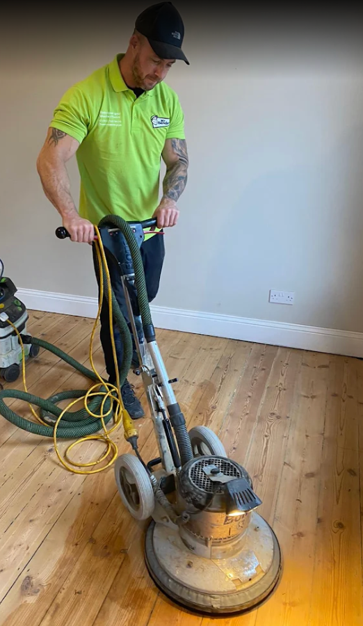 Man in green shirt sanding a wooden floor with a large machine, yellow hose, inside a room.