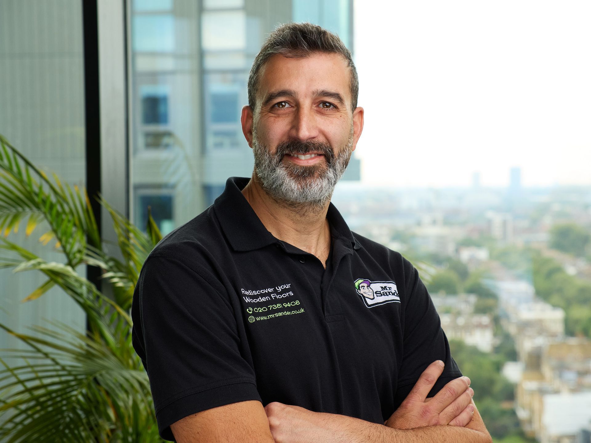 Man with a gray beard and black shirt, arms crossed, smiling at the camera, urban background.