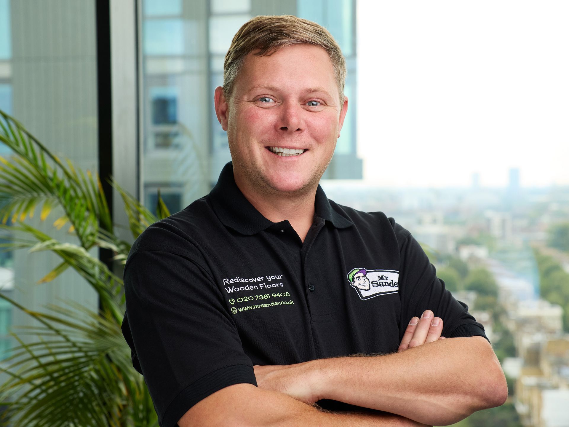 Man in black shirt with arms crossed, smiling, in front of window overlooking city with palm plant to the left.