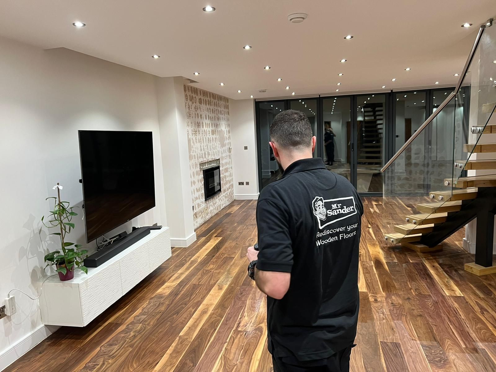 Man in black shirt surveys a modern living room with a TV, fireplace, and hardwood floors.