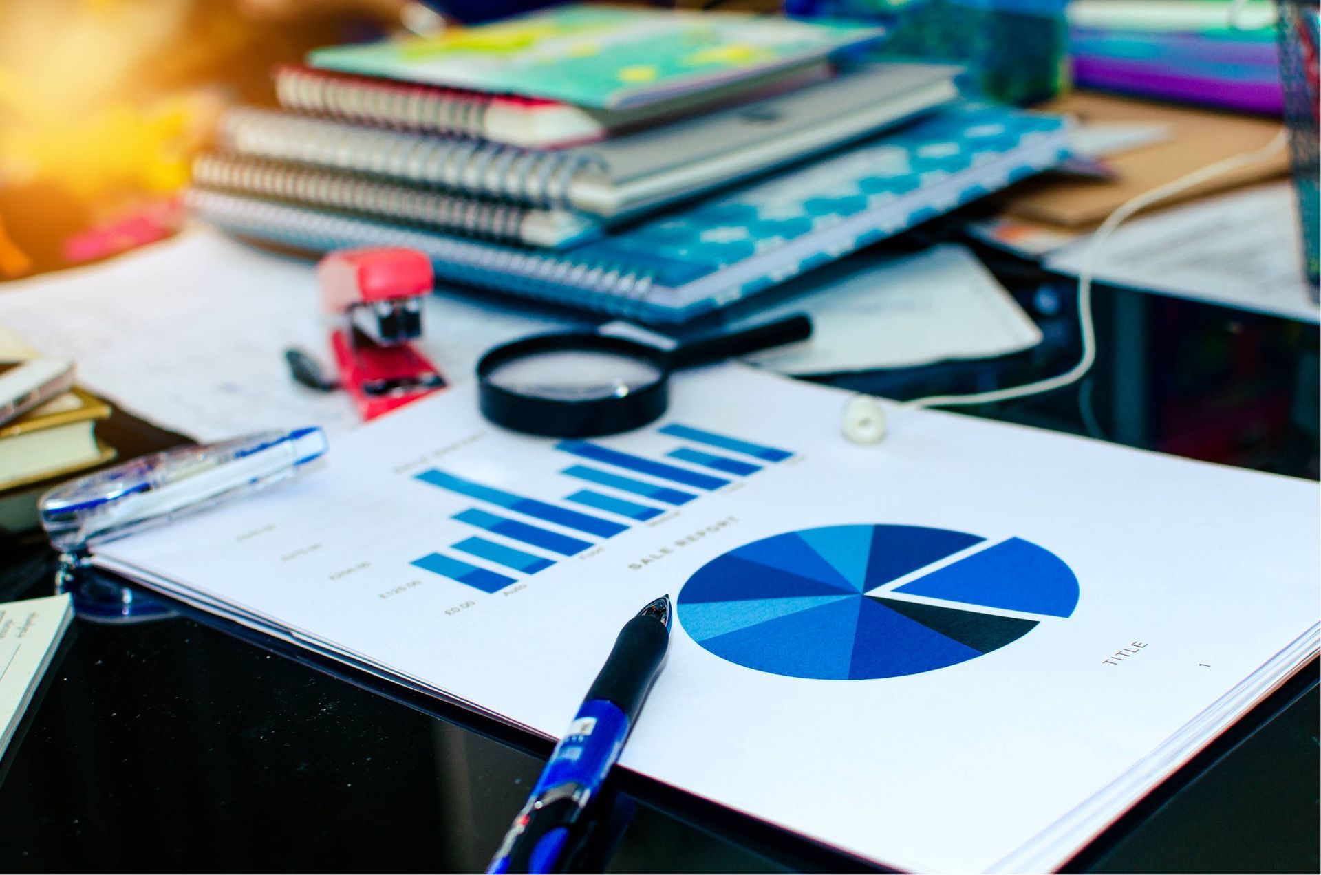 Financial graphs and charts with a pen, magnifying glass, stapler, and notebooks on a desk.