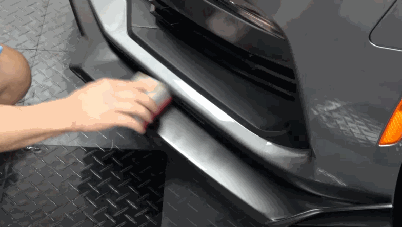 A technician applying ceramic coating to a car's front bumper, using a small applicator pad for precision.
