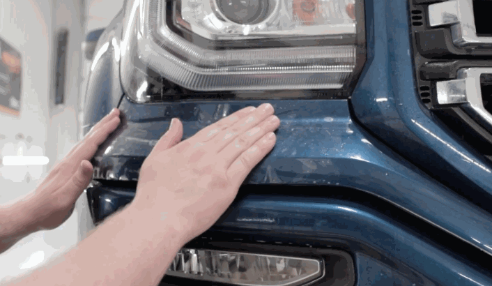 Hands smoothing out paint protection film on the front bumper of a blue truck near the headlight.